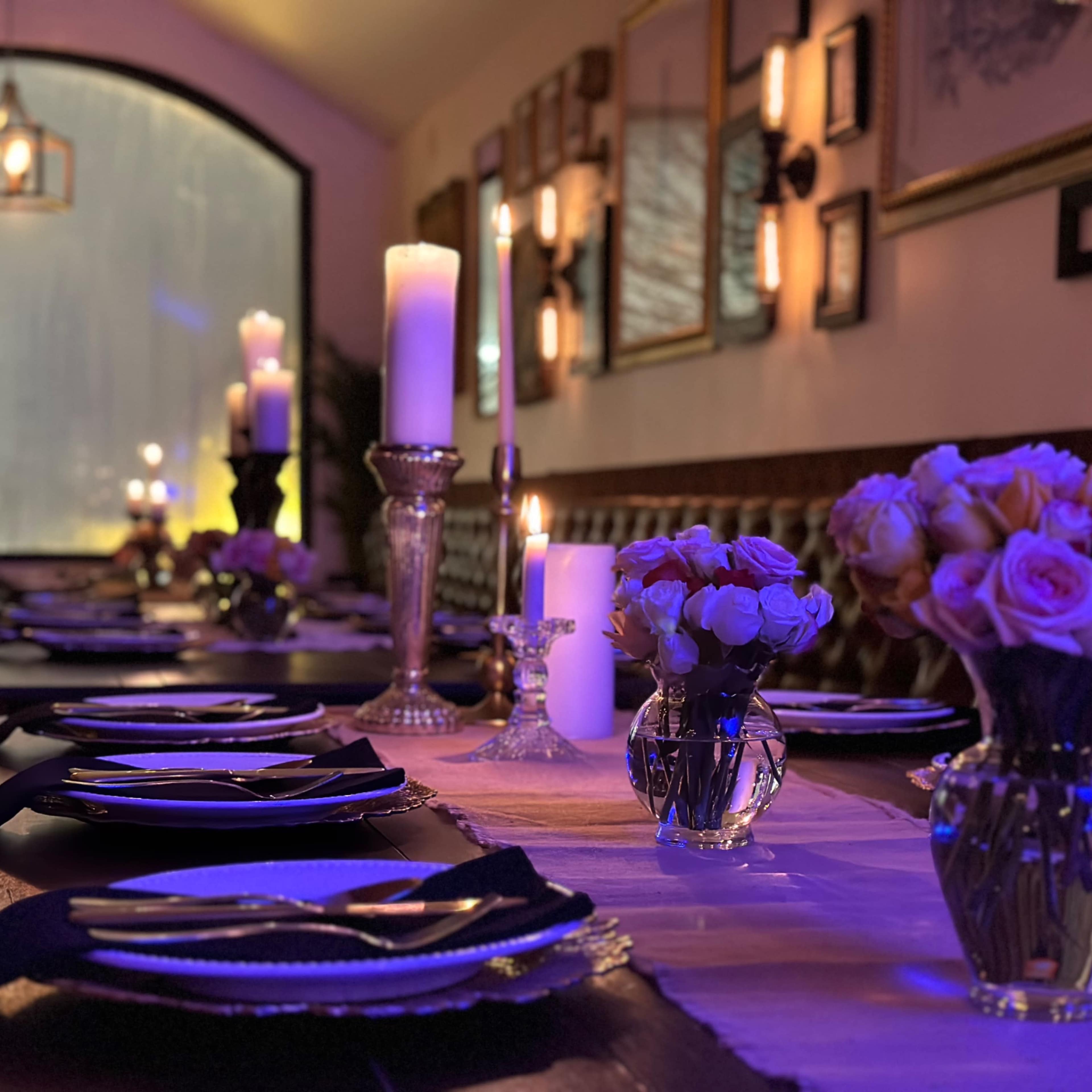 A dining table is elegantly set with plates, cutlery, and floral arrangements, surrounded by softly glowing candles in a warmly lit restaurant interior.
