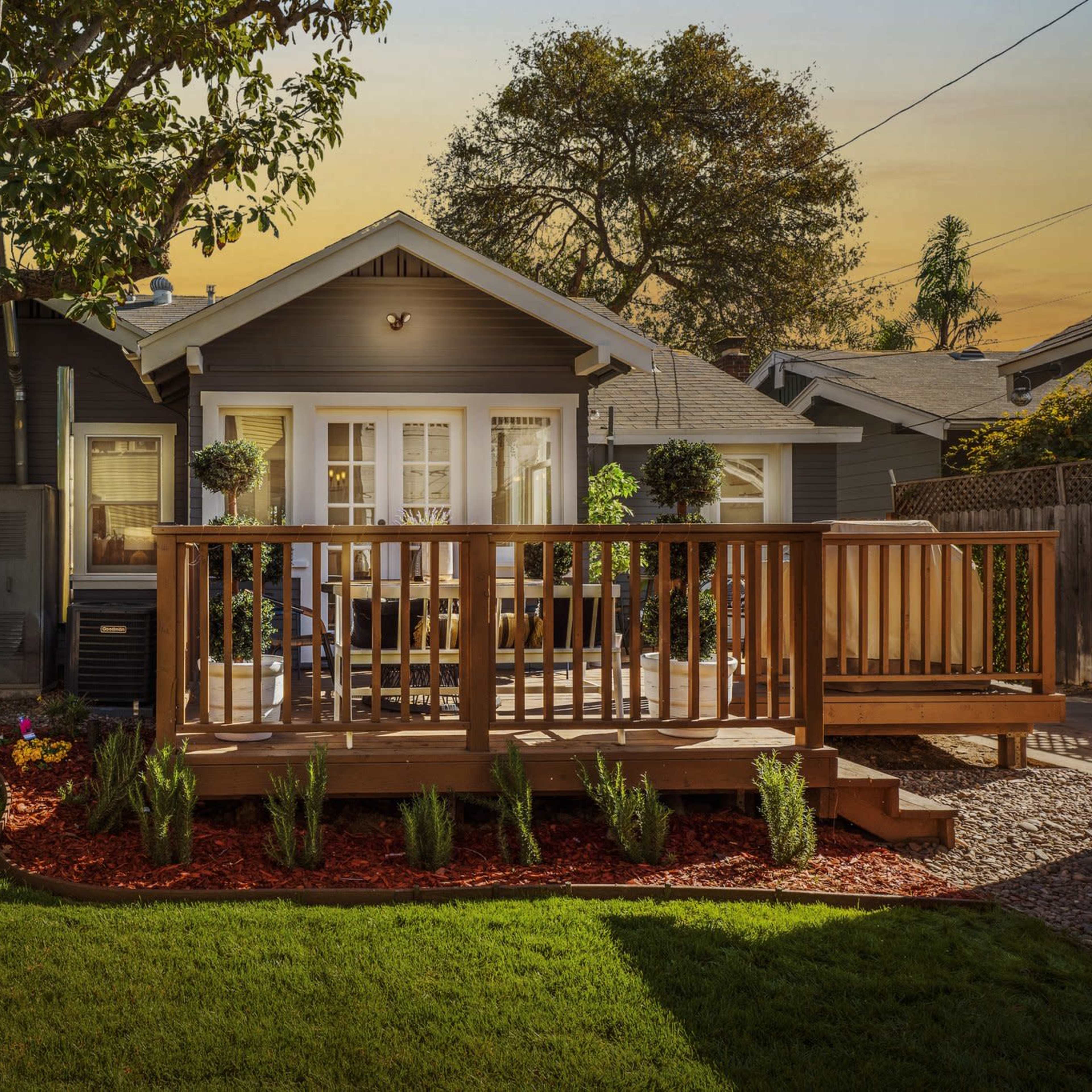 A house with a wooden deck is surrounded by a landscaped yard featuring grass, flowers, and trees in the golden light of sunset.