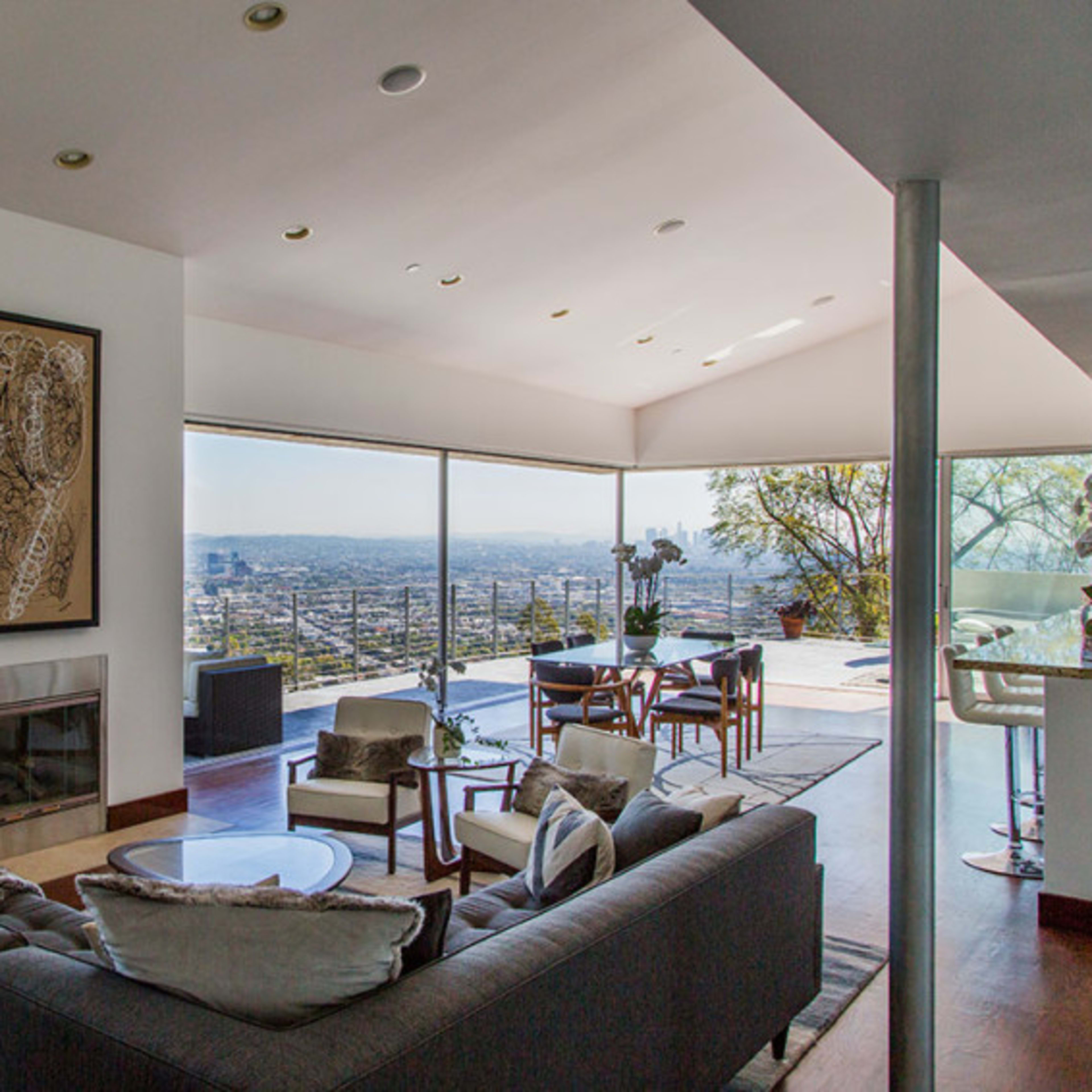 A modern living room with large windows offering a view of the city skyline, featuring a gray sofa, a glass coffee table, and a dining area in the background.
