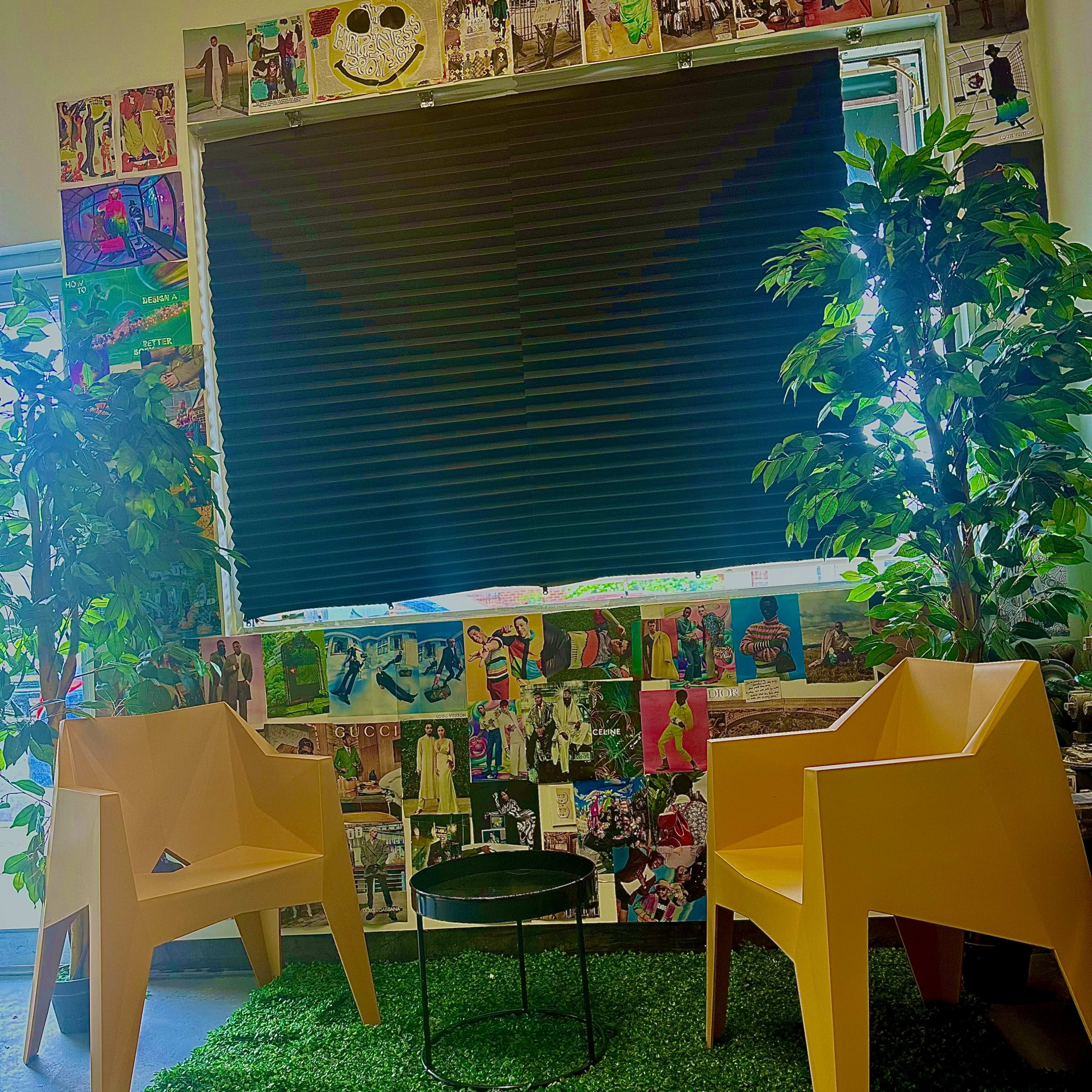 The scene features two yellow chairs and a small black table on green artificial grass, set against a backdrop of a window covered by black shades and a wall adorned with vibrant collages.