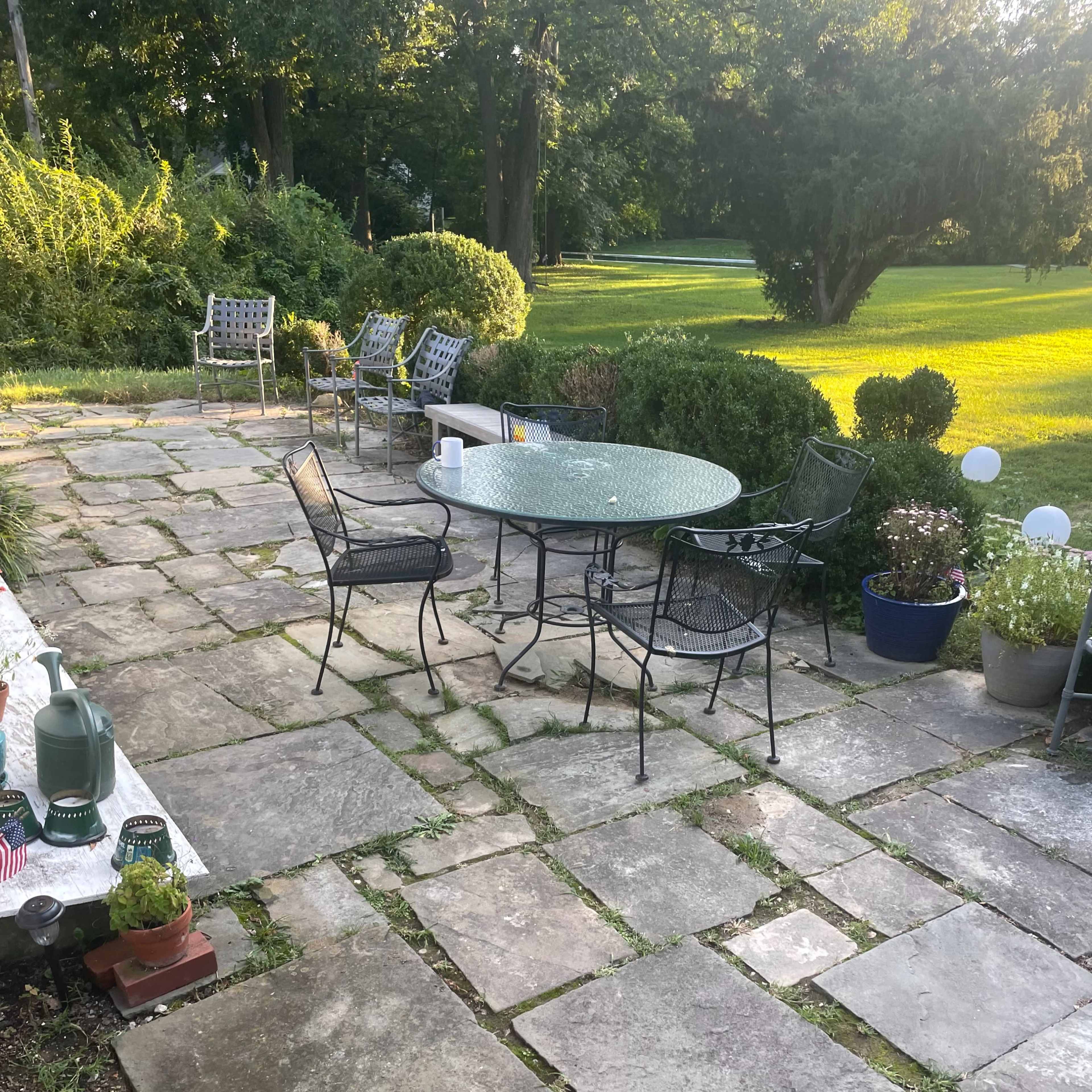 A stone patio with a round table and several chairs surrounded by greenery and potted plants.
