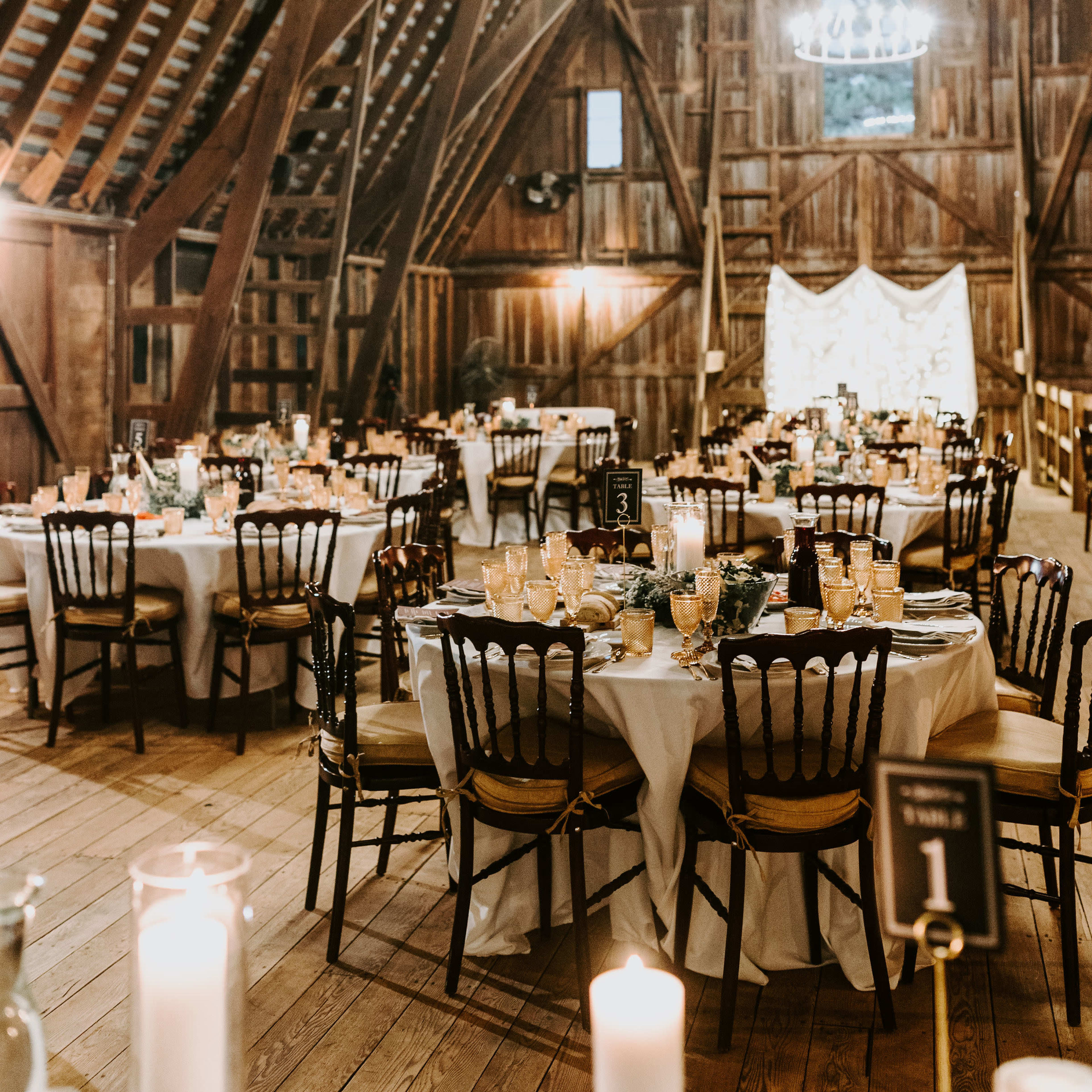 The interior of a rustic barn is set up for a banquet, featuring tables adorned with white linens and centerpieces, surrounded by wooden beams and warm lighting.