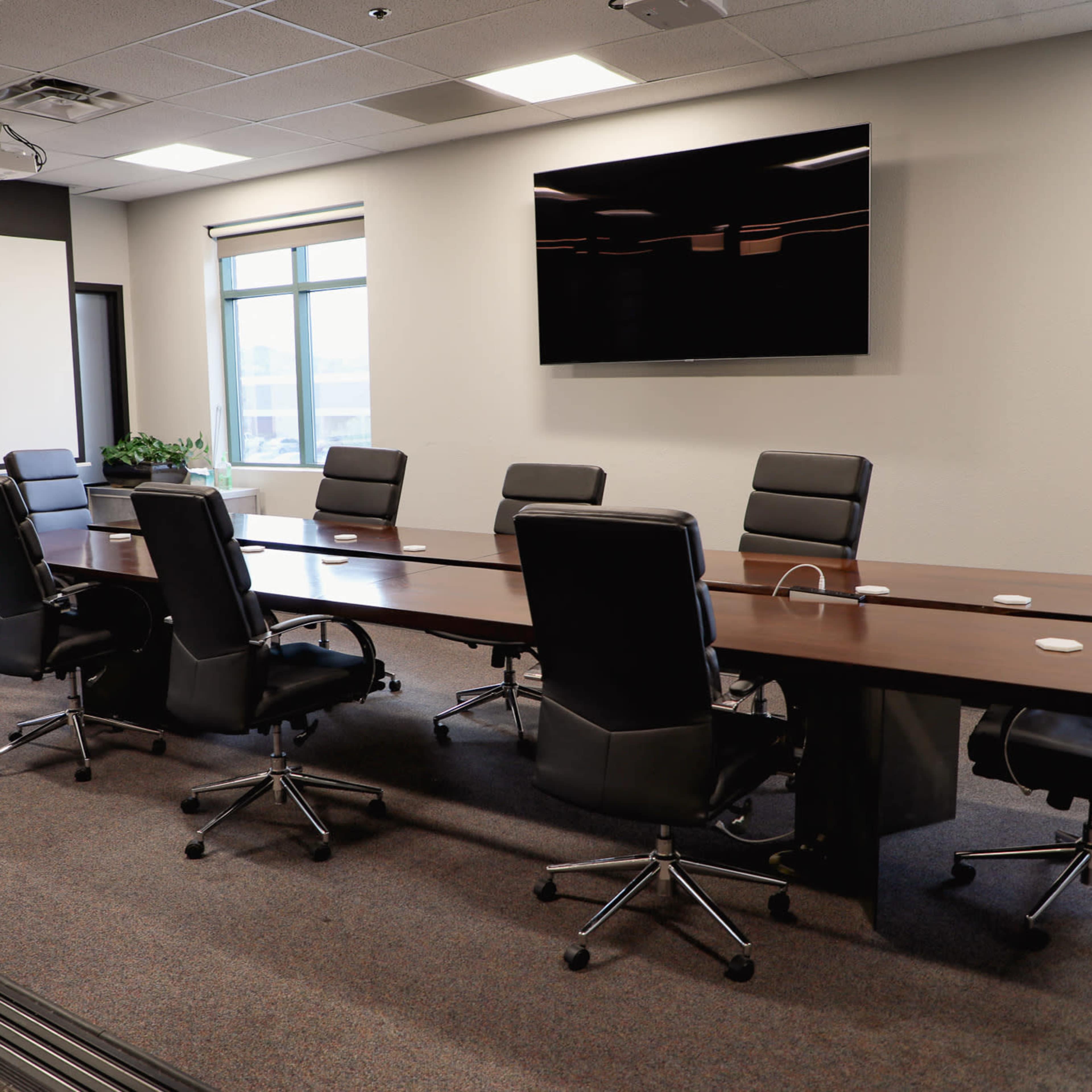 A modern conference room features a long wooden table surrounded by black rolling chairs, with a large screen mounted on the wall and windows allowing natural light.