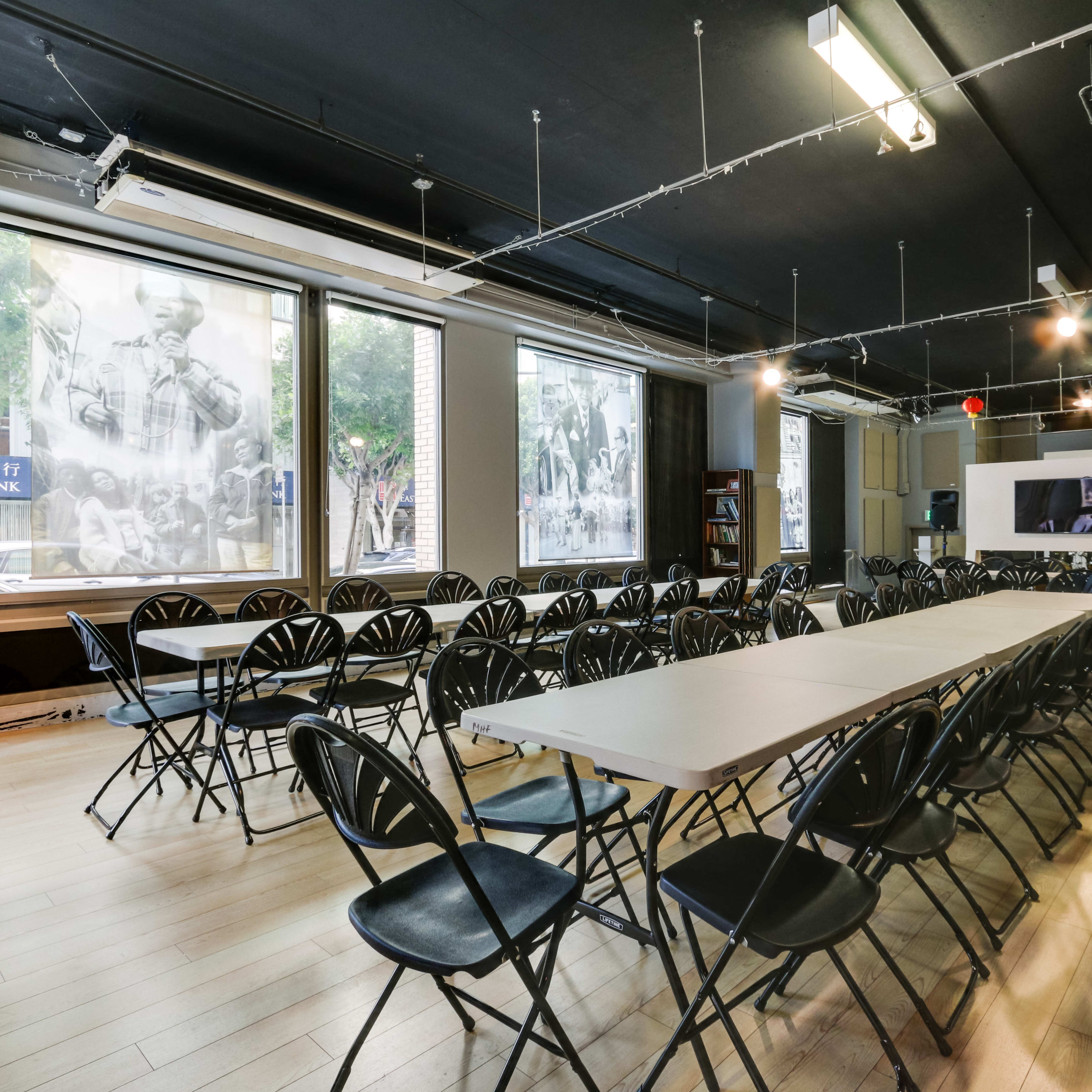 A spacious room is set up with rows of folding chairs facing a long table, surrounded by large windows showcasing black-and-white photographs.