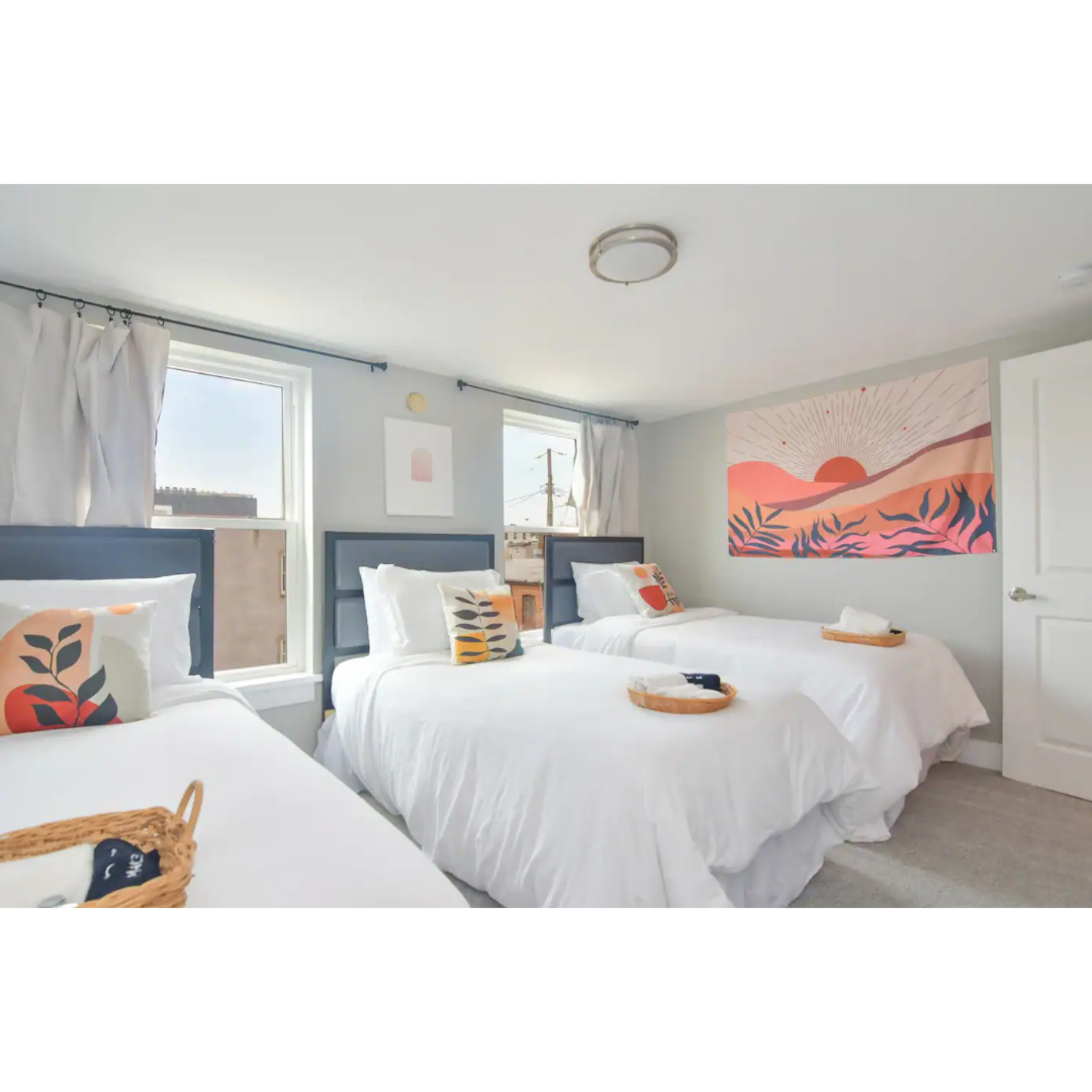 A bright bedroom features three beds with white linens, a large wall mural of a sunset, and windows allowing natural light.