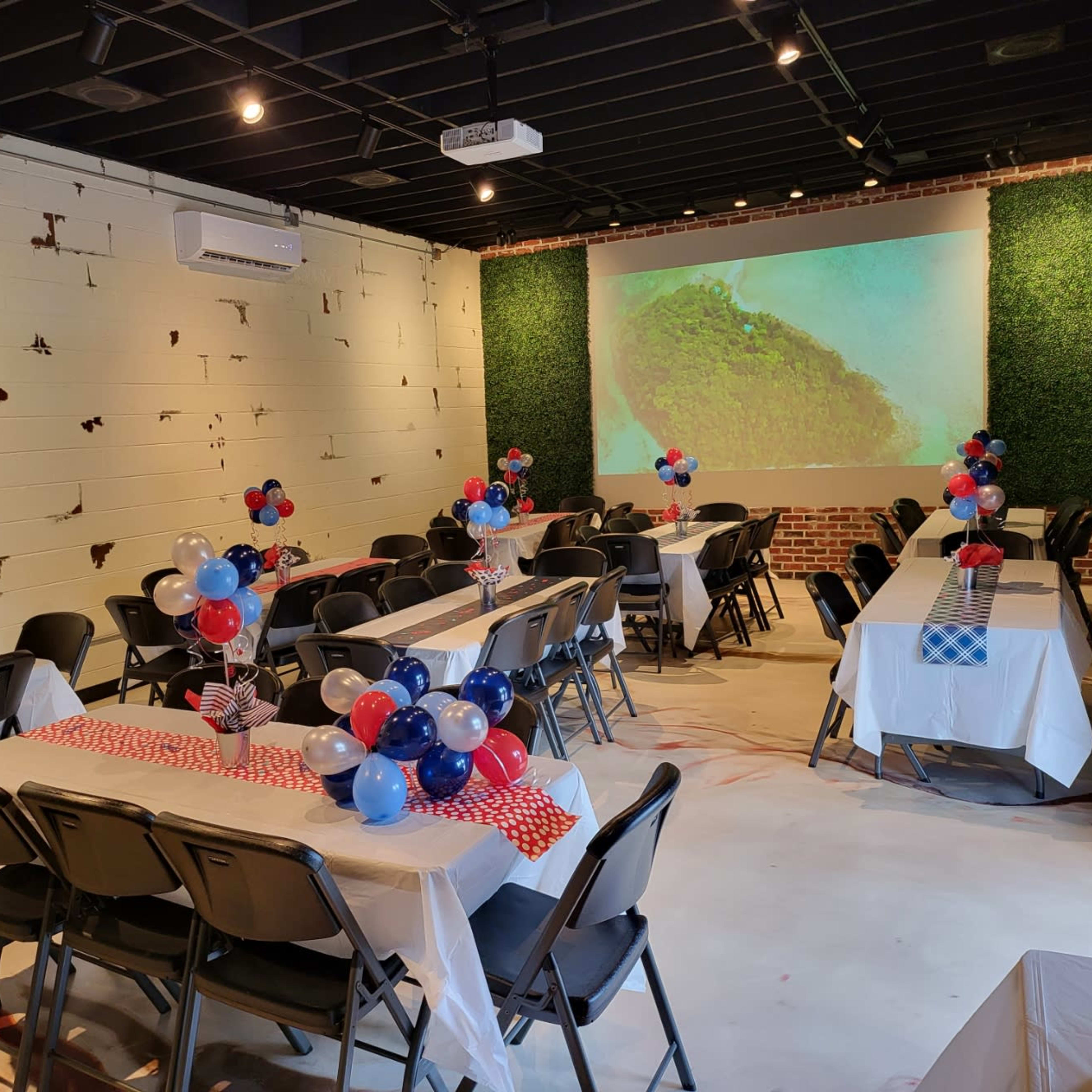 A decorated indoor event space with tables set up in rows, featuring patriotic balloons and tablecloths, and a large screen displaying a scenic image at the front.