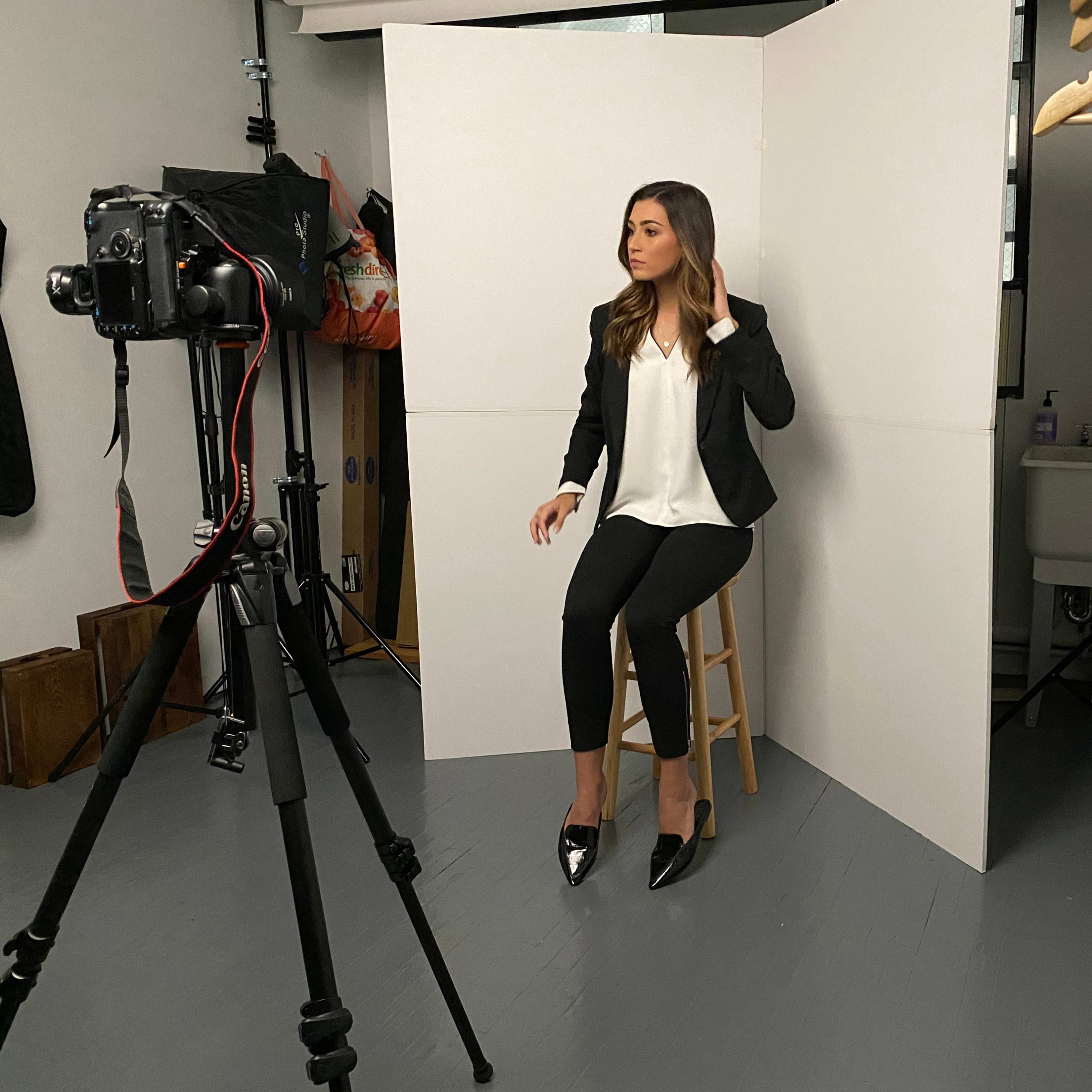 A woman in a black blazer and high heels is sitting on a stool in a photography studio, posing for the camera.