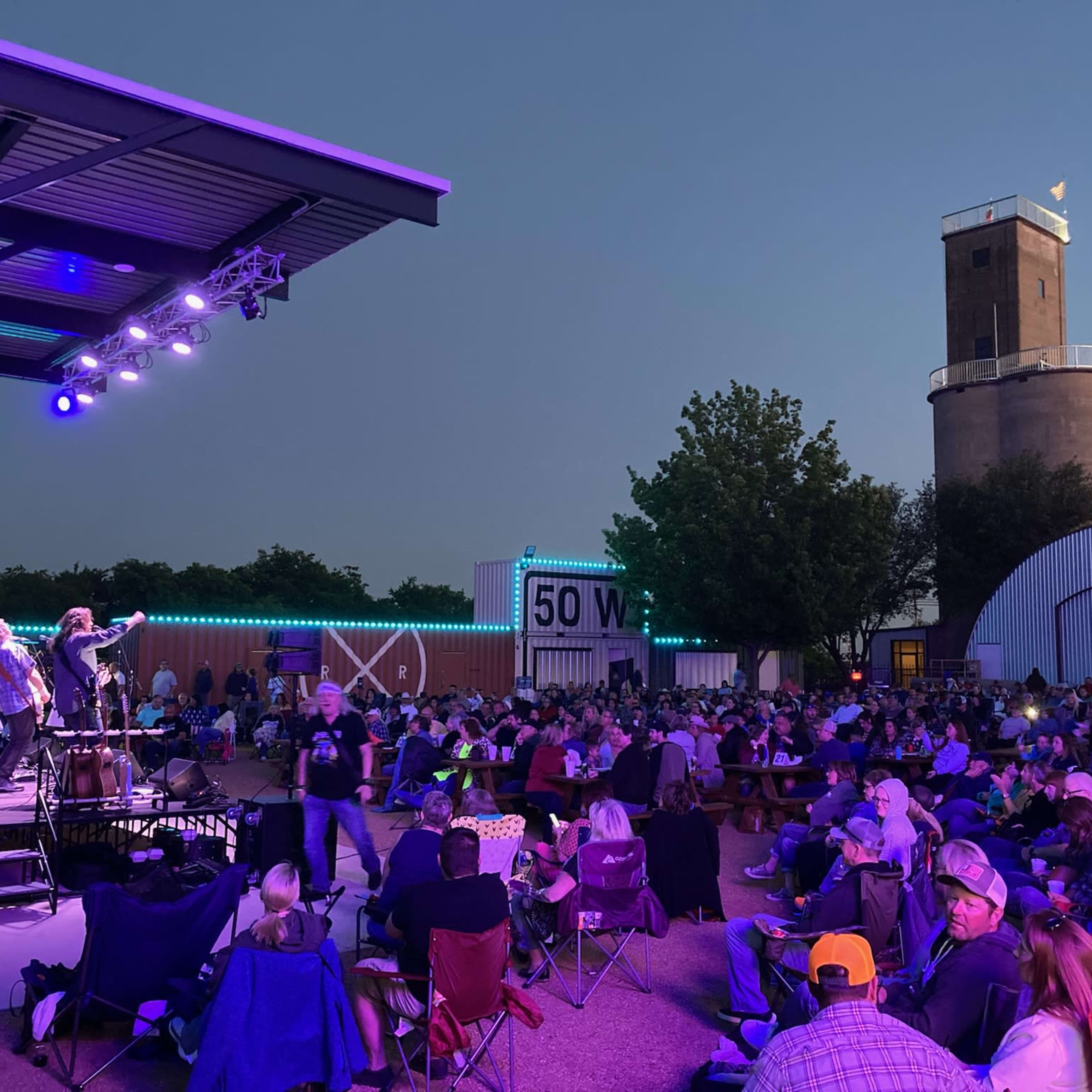 A large crowd sits in front of a stage, enjoying a live music performance at dusk, with a tower and buildings in the background.