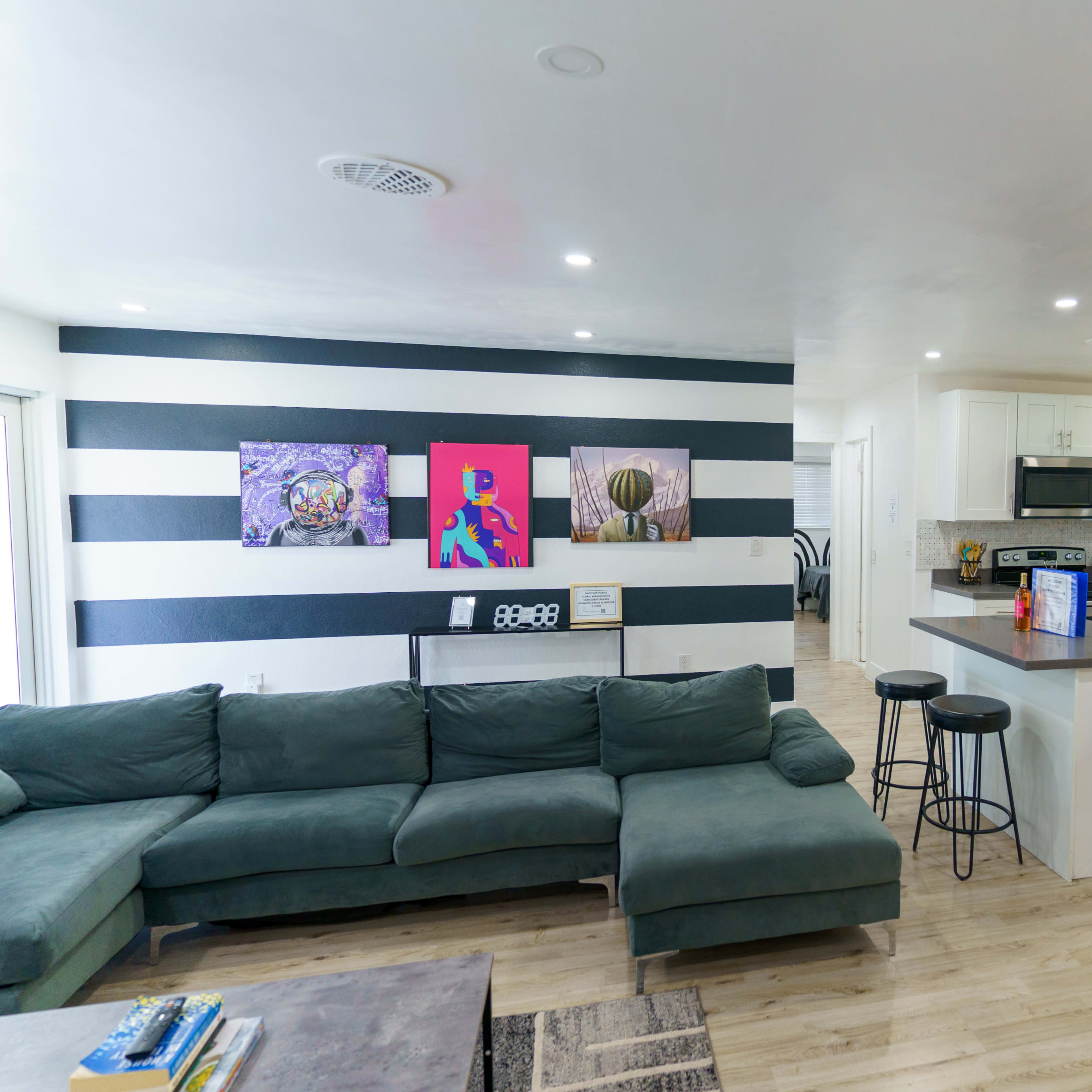 A modern open-concept living area features a green sectional sofa, black bar stools, and a kitchen with white cabinets and stainless steel appliances, complemented by colorful wall art.