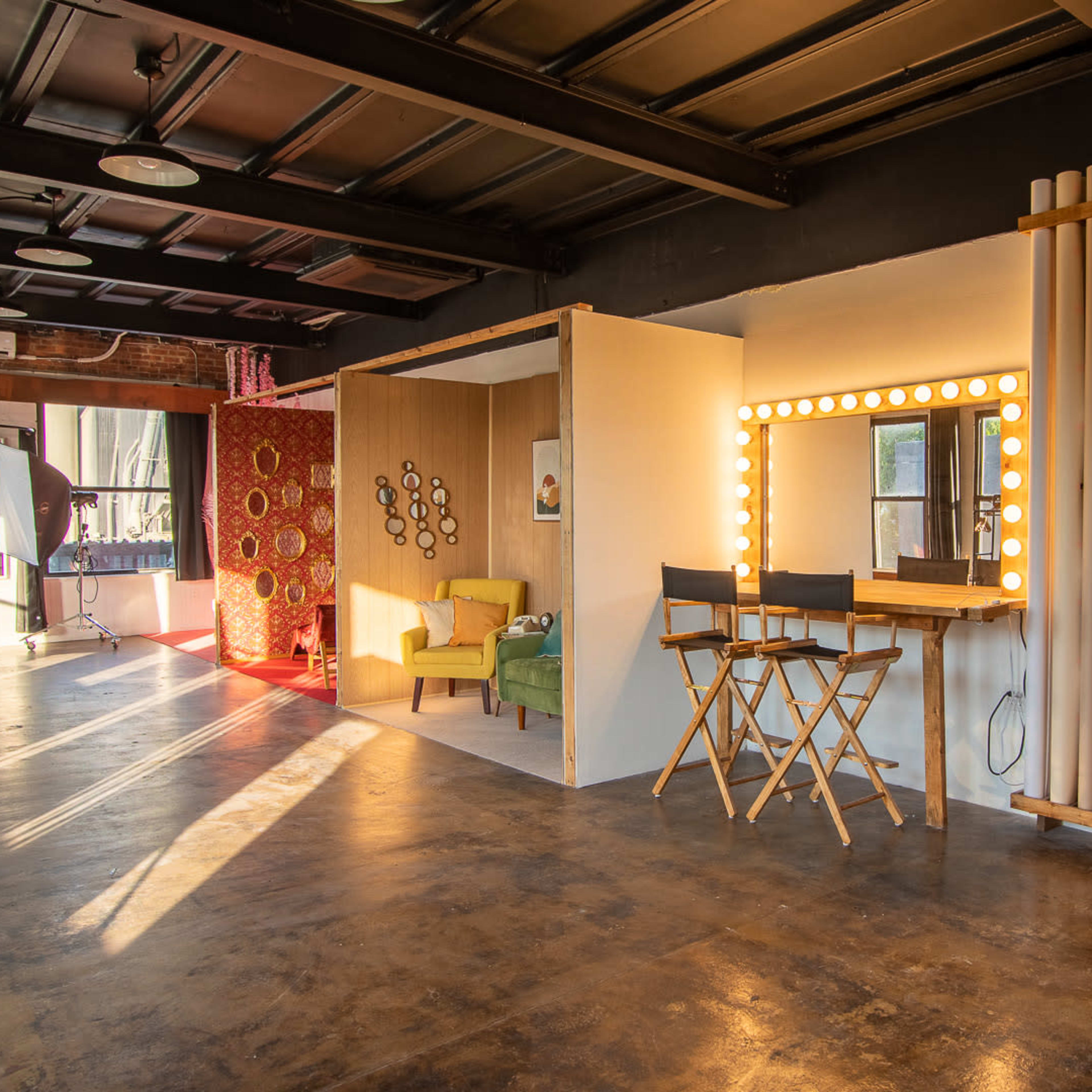 A well-lit studio space featuring various backdrops, a makeup mirror with lights, and director's chairs arranged in an organized layout.