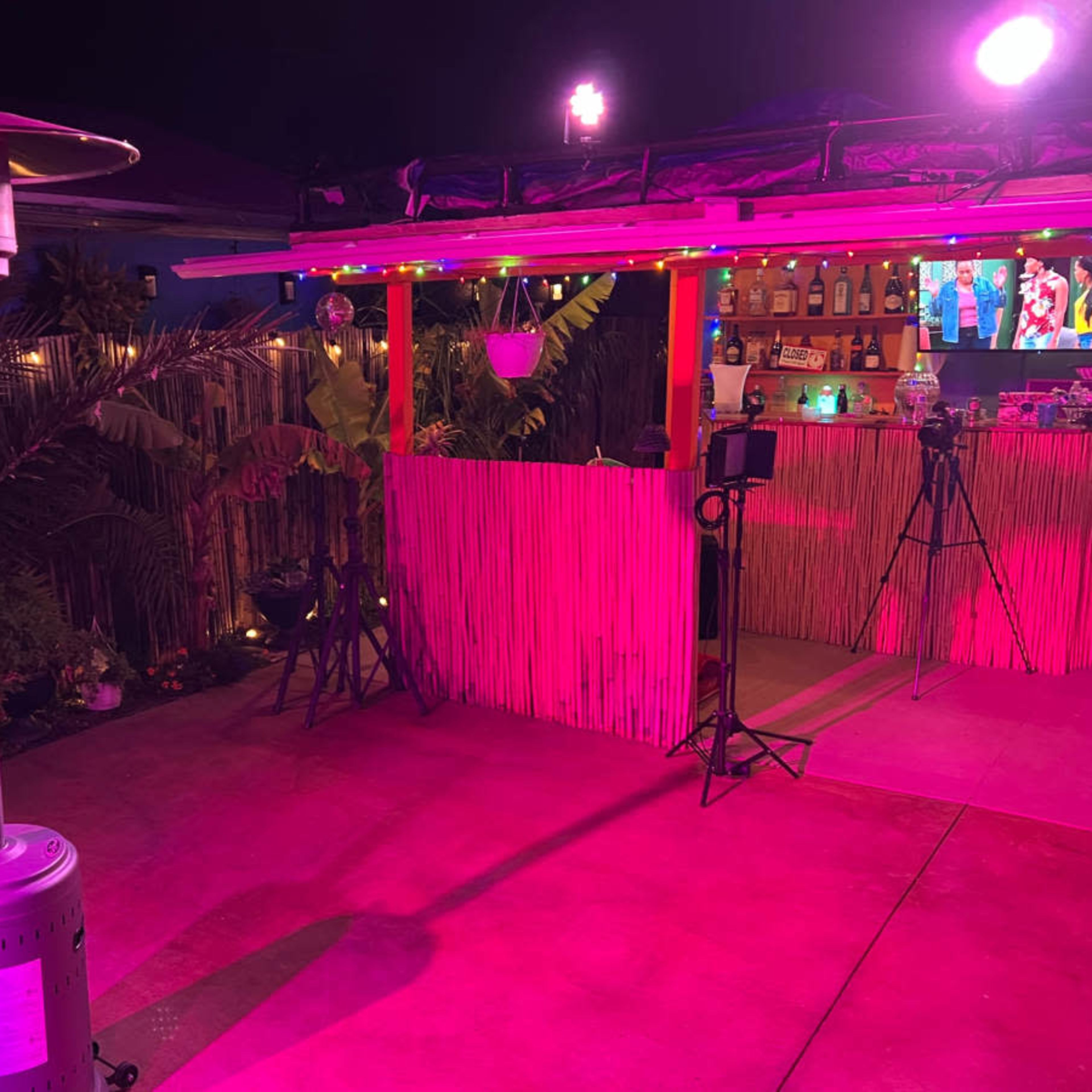 A brightly lit outdoor space with a patio heater, colorful lights, and a bar area surrounded by tropical plants.