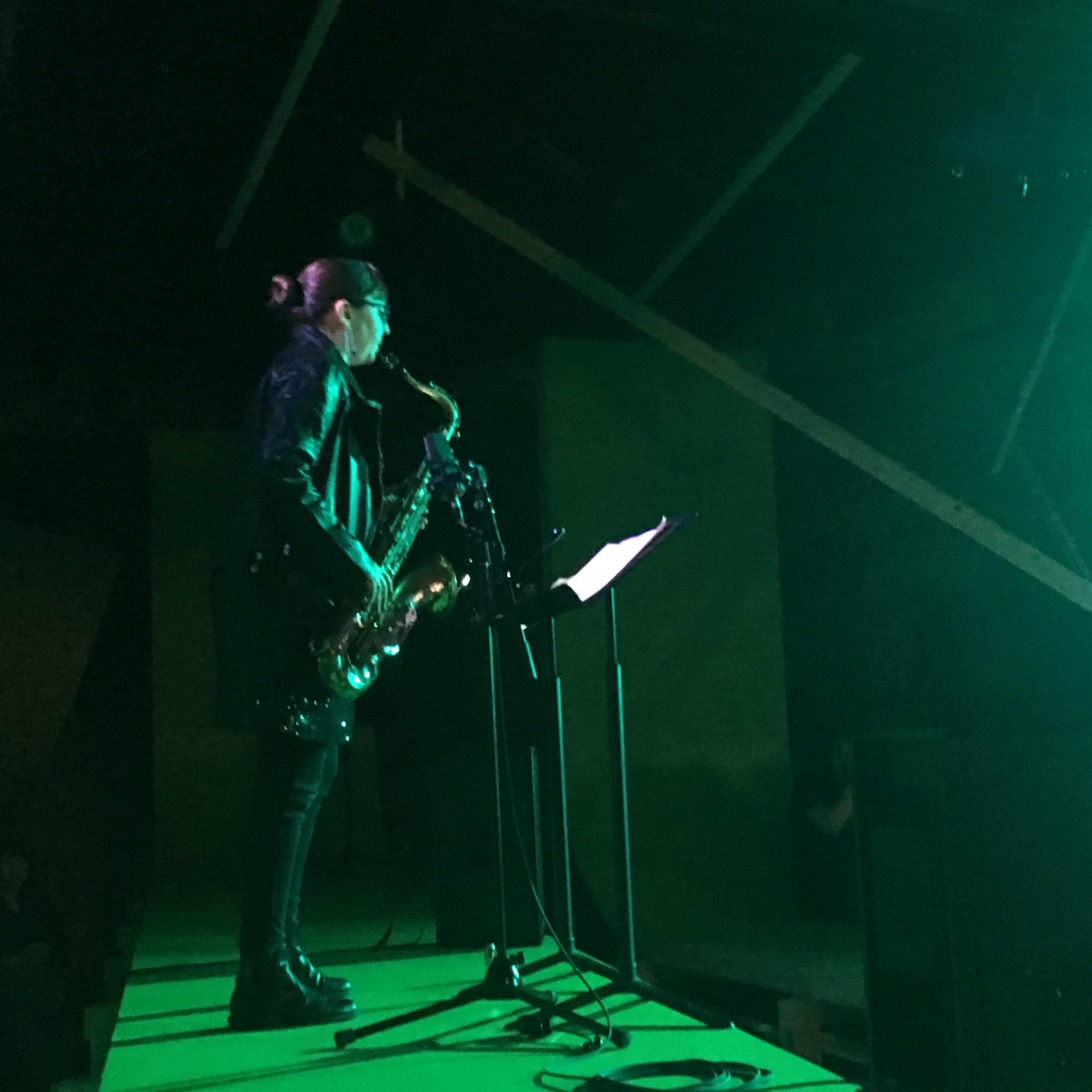 A musician plays the saxophone on a stage illuminated by green lighting, with audience members visible in the background.