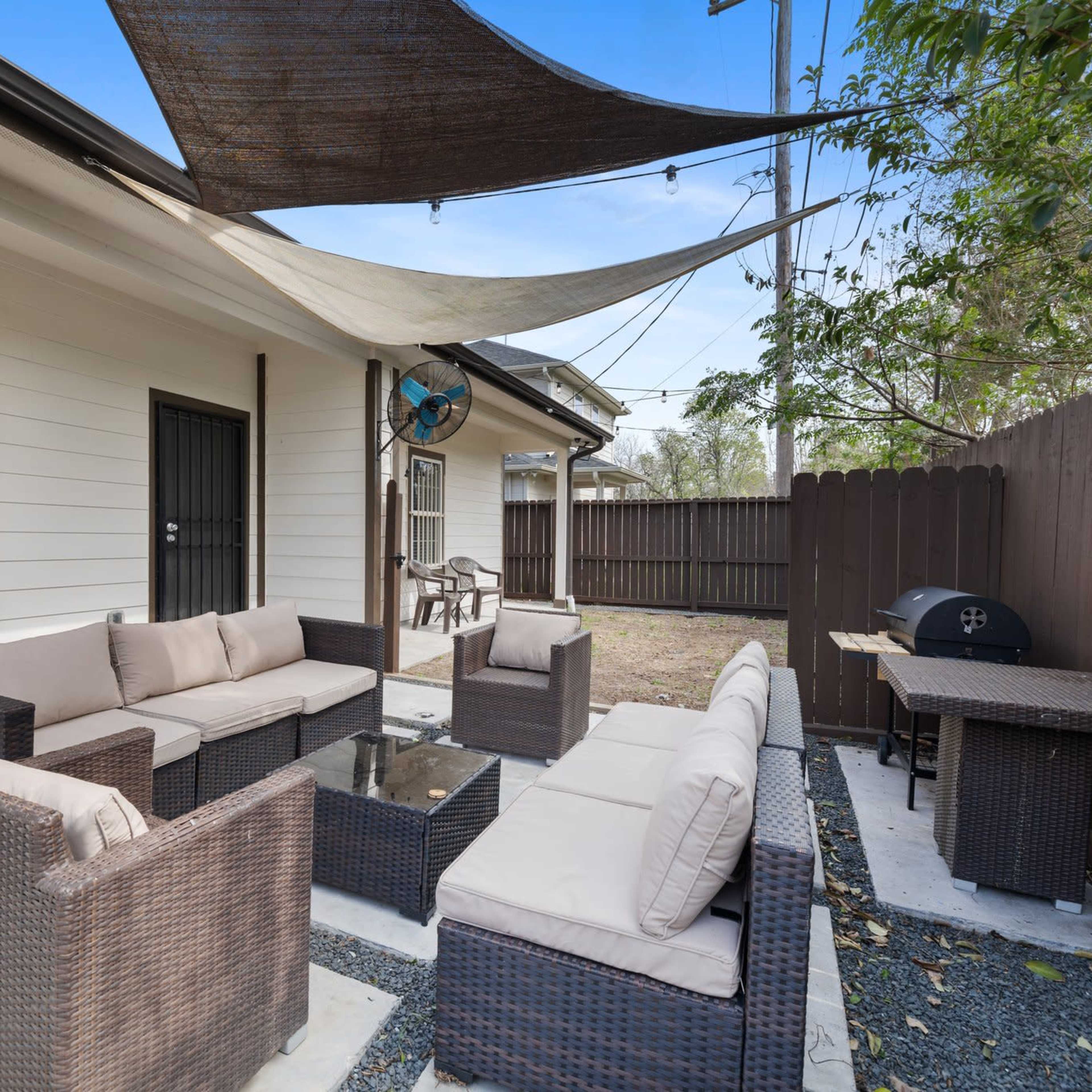 A covered outdoor seating area with a table surrounded by wicker furniture and a barbecue grill on a stone path beside a wooden fence.