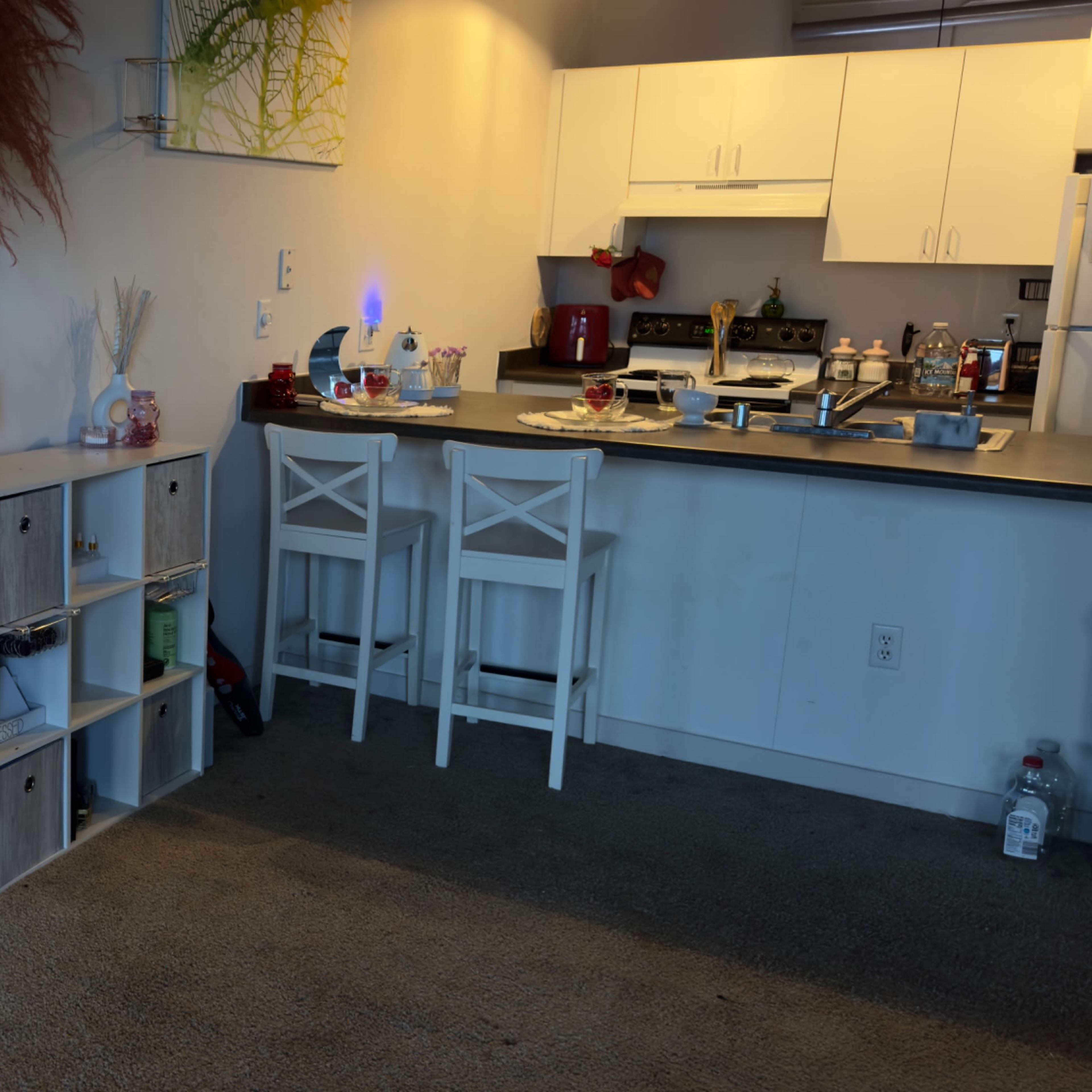 A furnished living area with a large mirror, a white storage unit, and a kitchen counter with stools in a well-lit space.