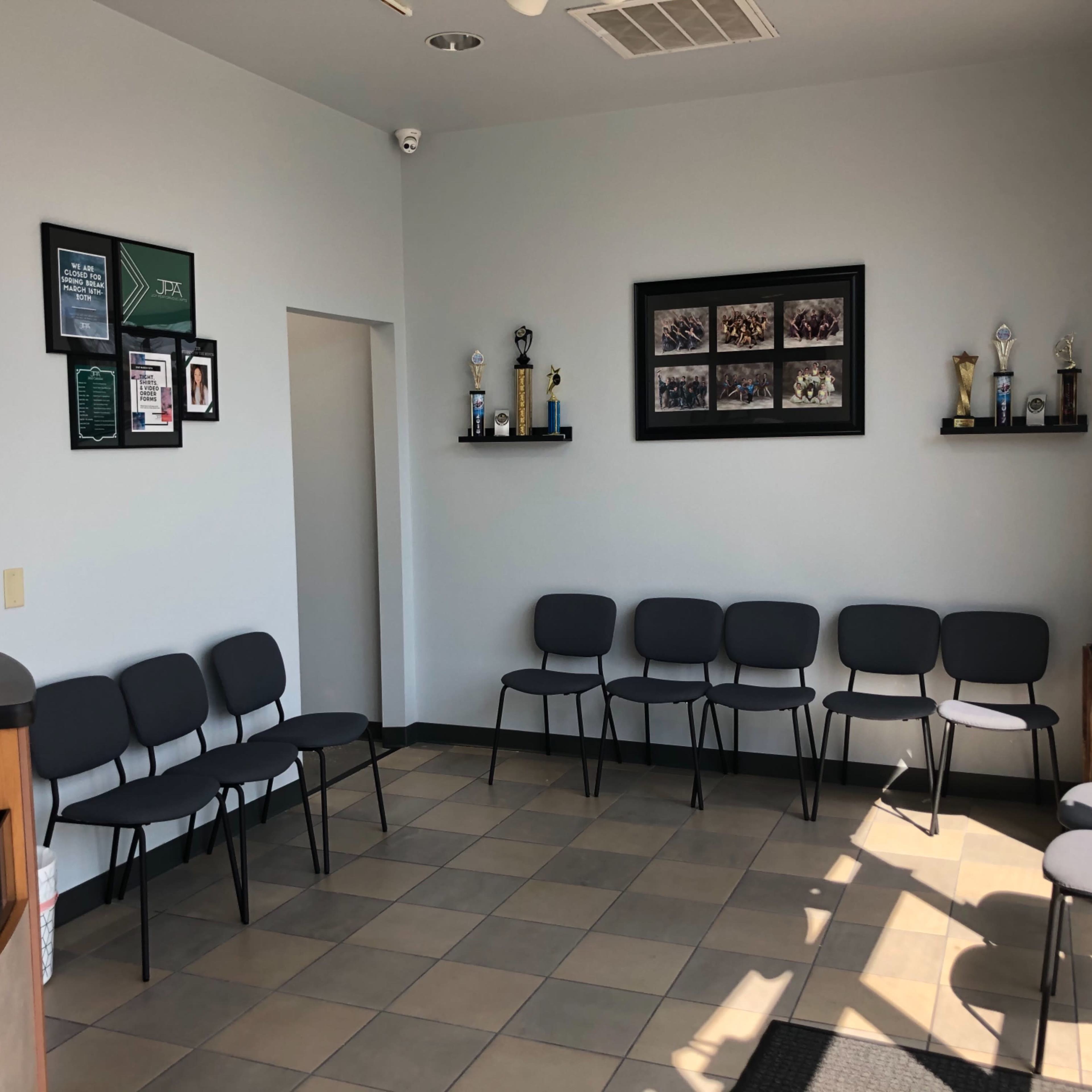 A waiting area with a reception desk, several black chairs arranged in a row, and a wall displaying trophies and framed photographs.