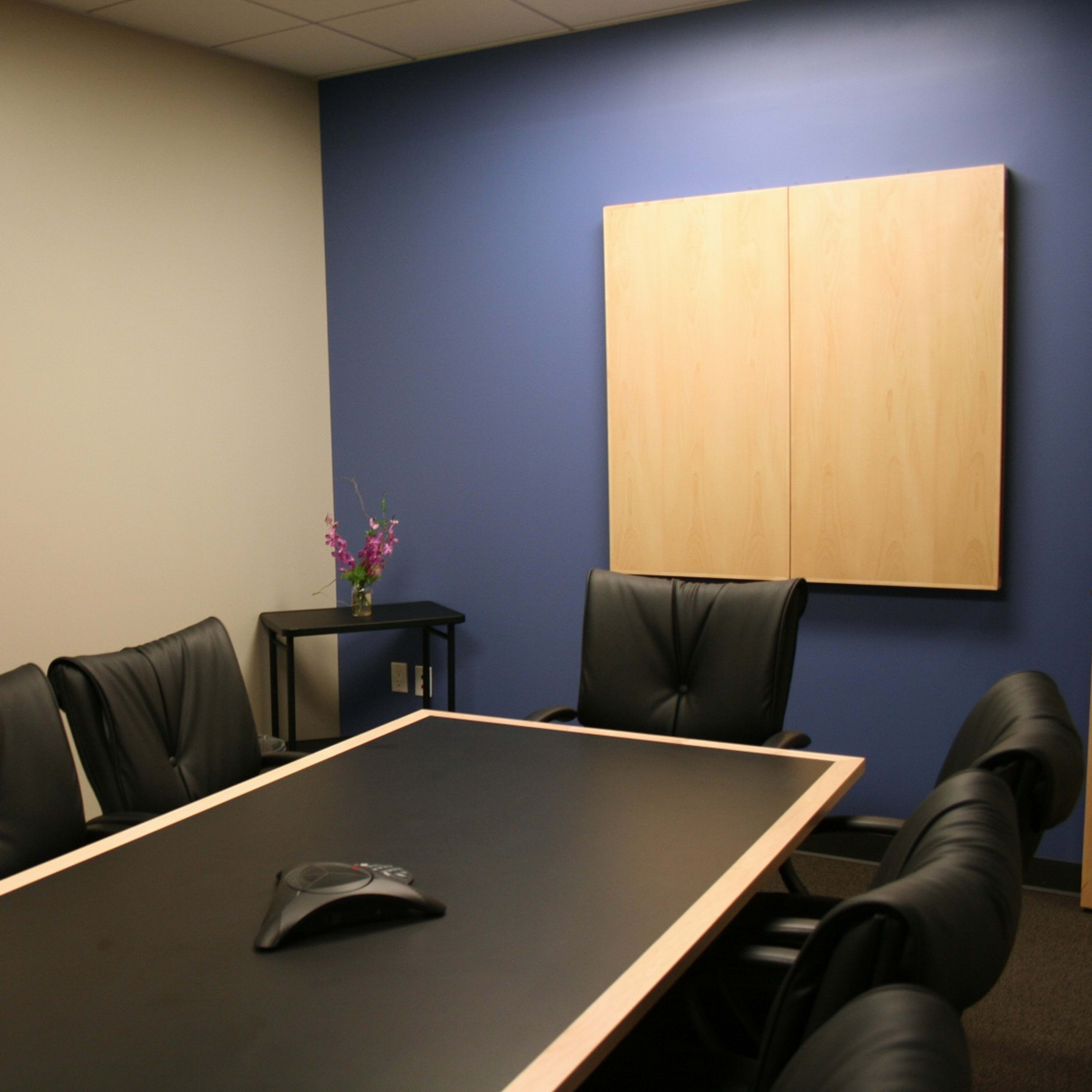 A modern conference room features a long black table surrounded by black leather chairs, with a landscape painting on one wall and a wooden cabinet on another.