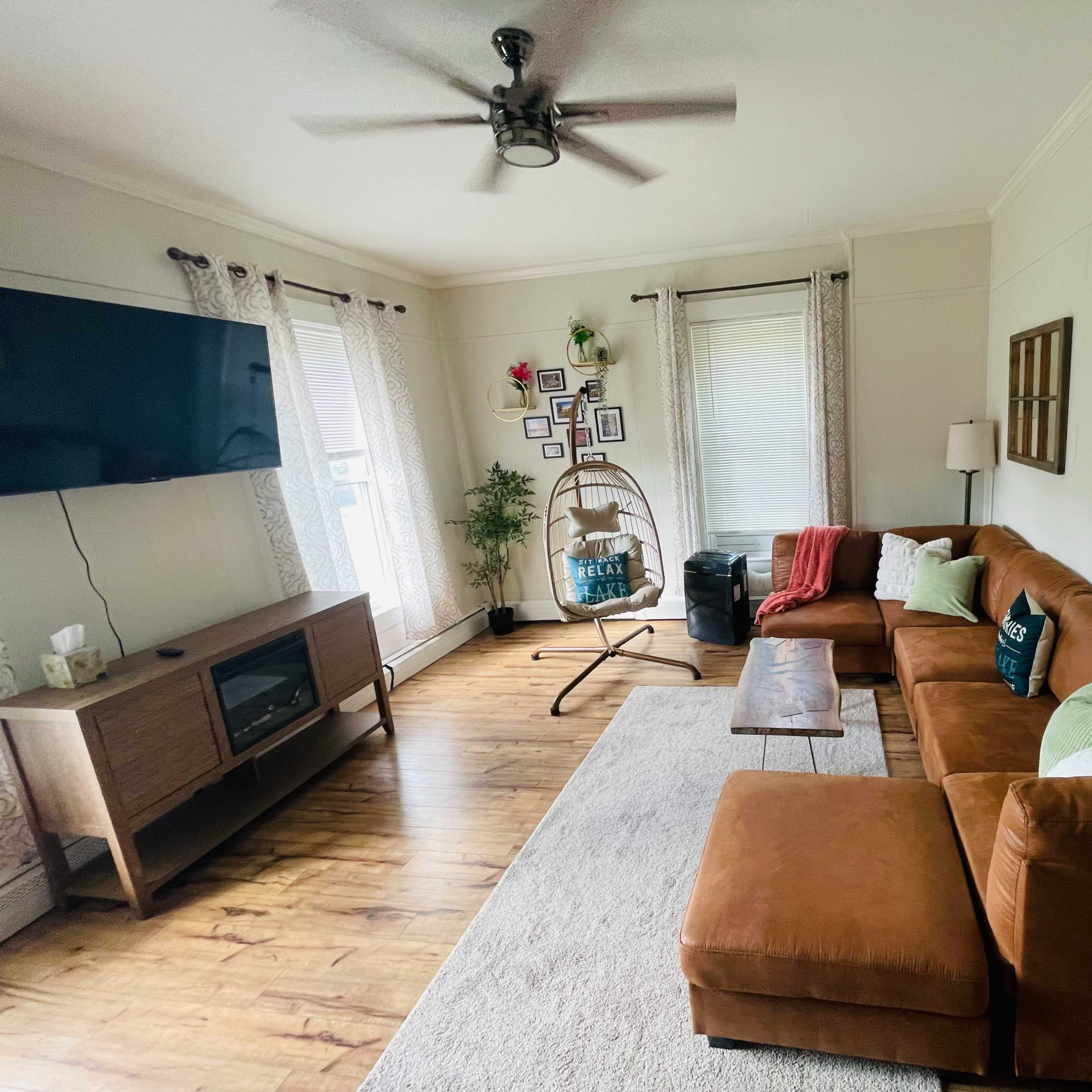 A cozy living room with a brown sectional sofa, a wooden TV stand, and large windows covered with light curtains.