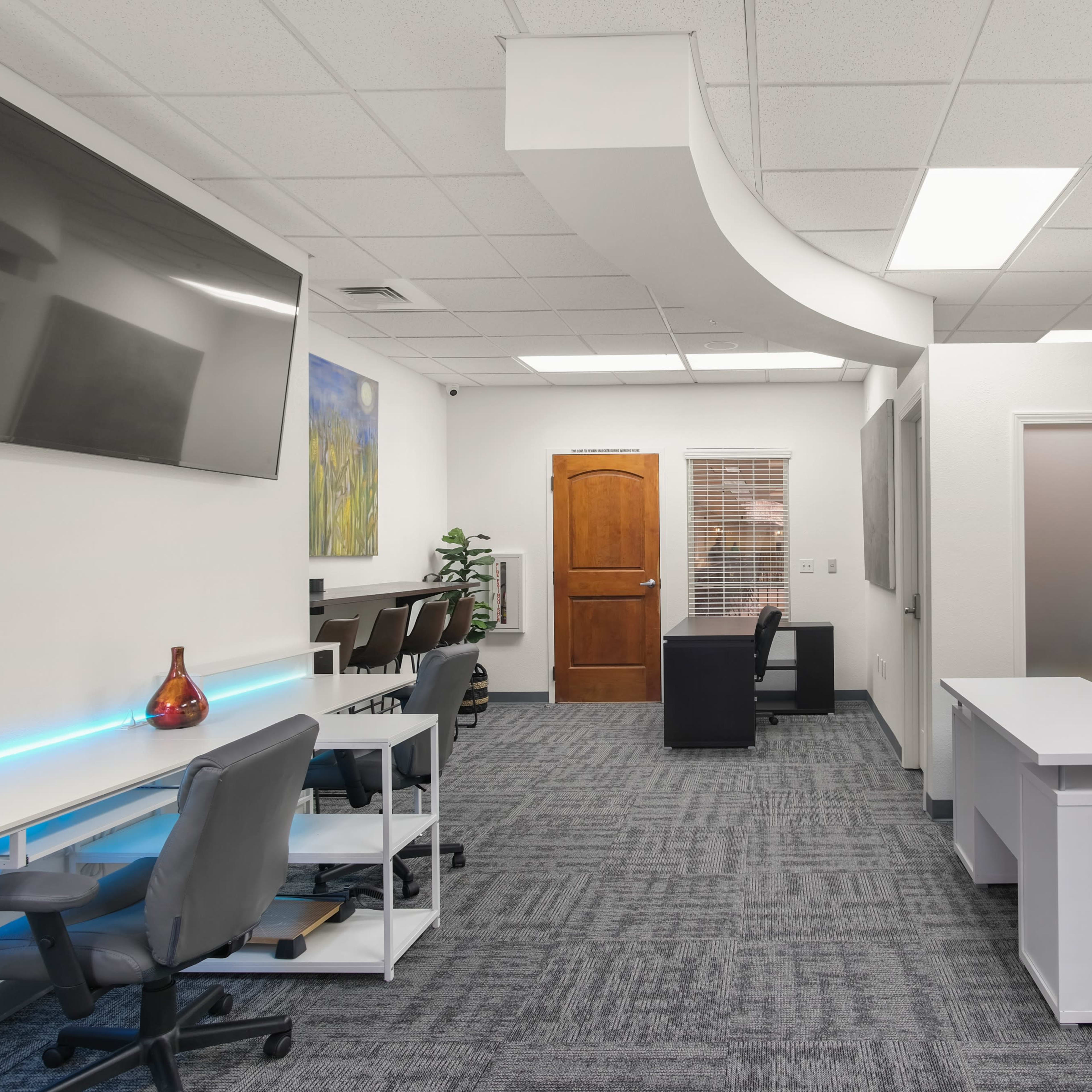 A modern office space with multiple workstations, a wooden door, and a large television mounted on the wall.