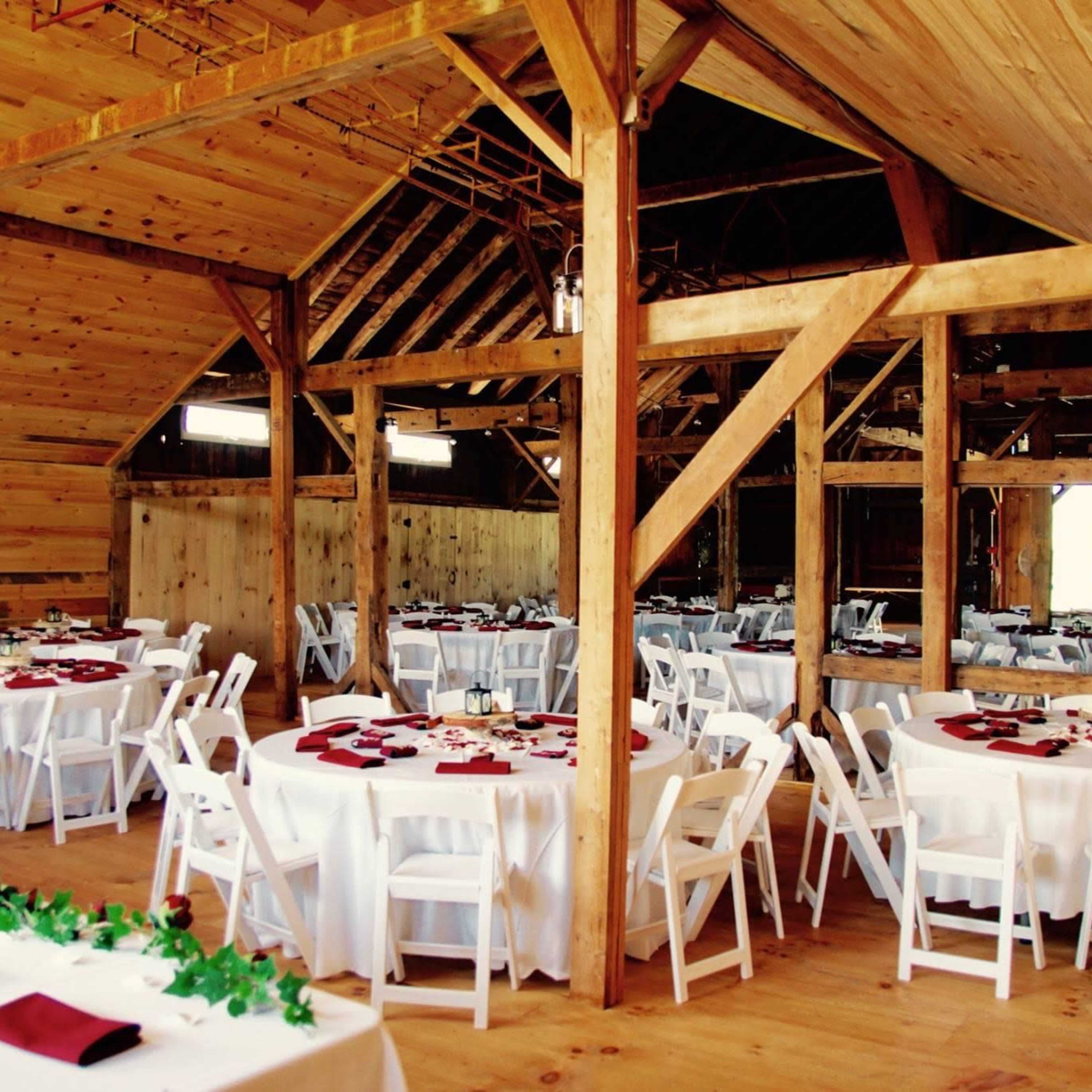 A spacious barn interior set up for an event, featuring several round tables with white tablecloths and red napkins.