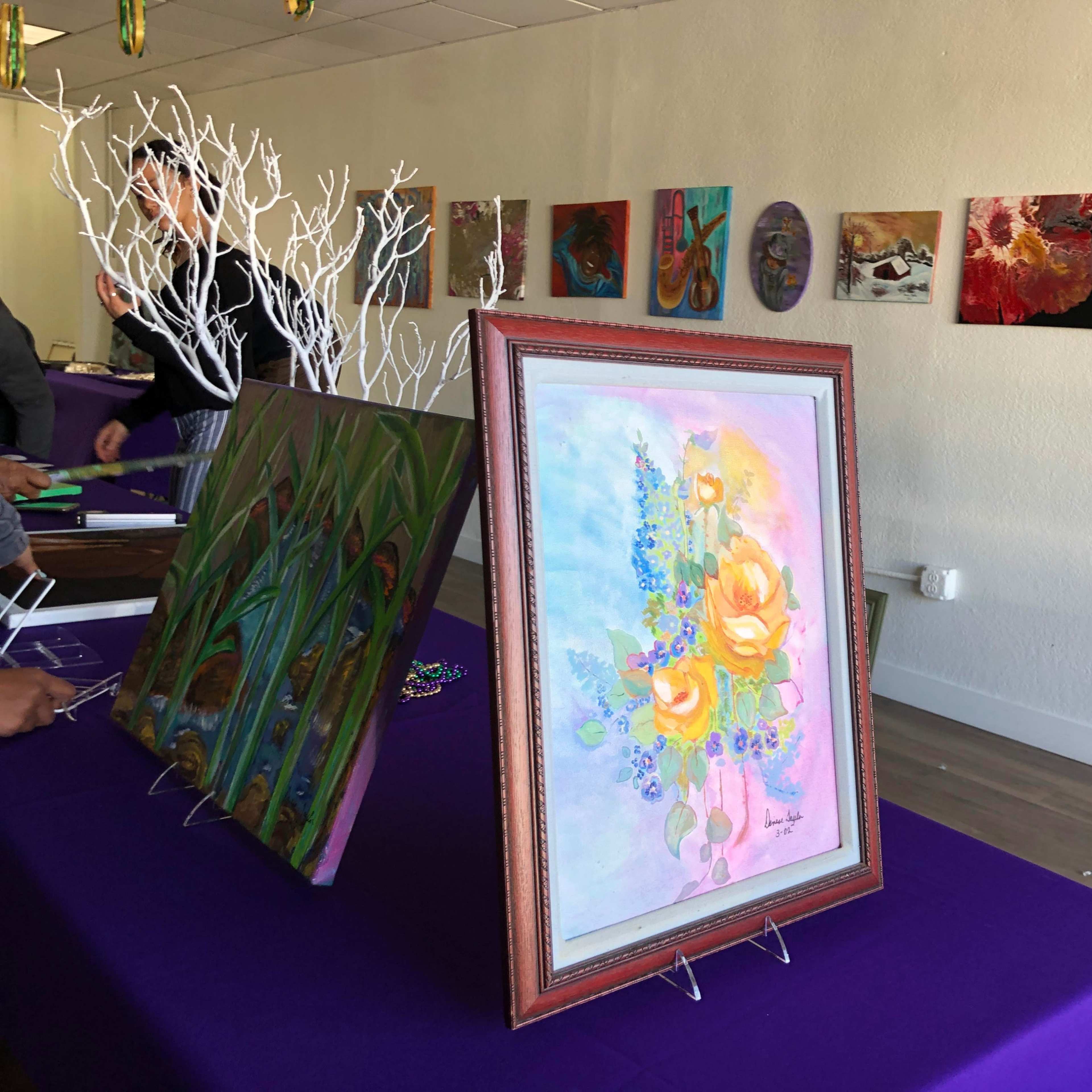 A gallery setting with various paintings displayed on the walls, and a framed floral painting in the foreground set against a purple tablecloth.