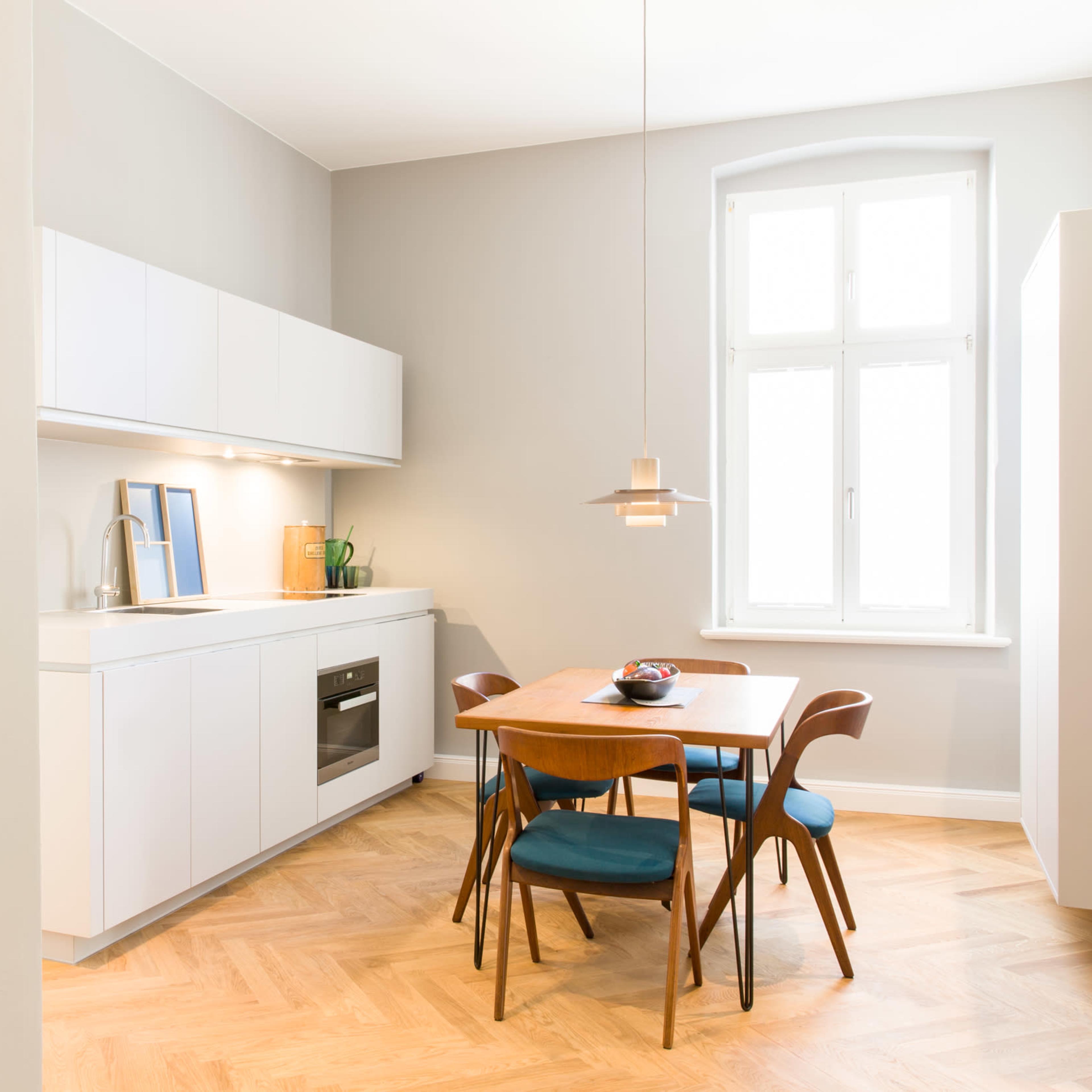 A modern kitchen with a wooden dining table, four chairs, sleek white cabinetry, and large windows allowing natural light.
