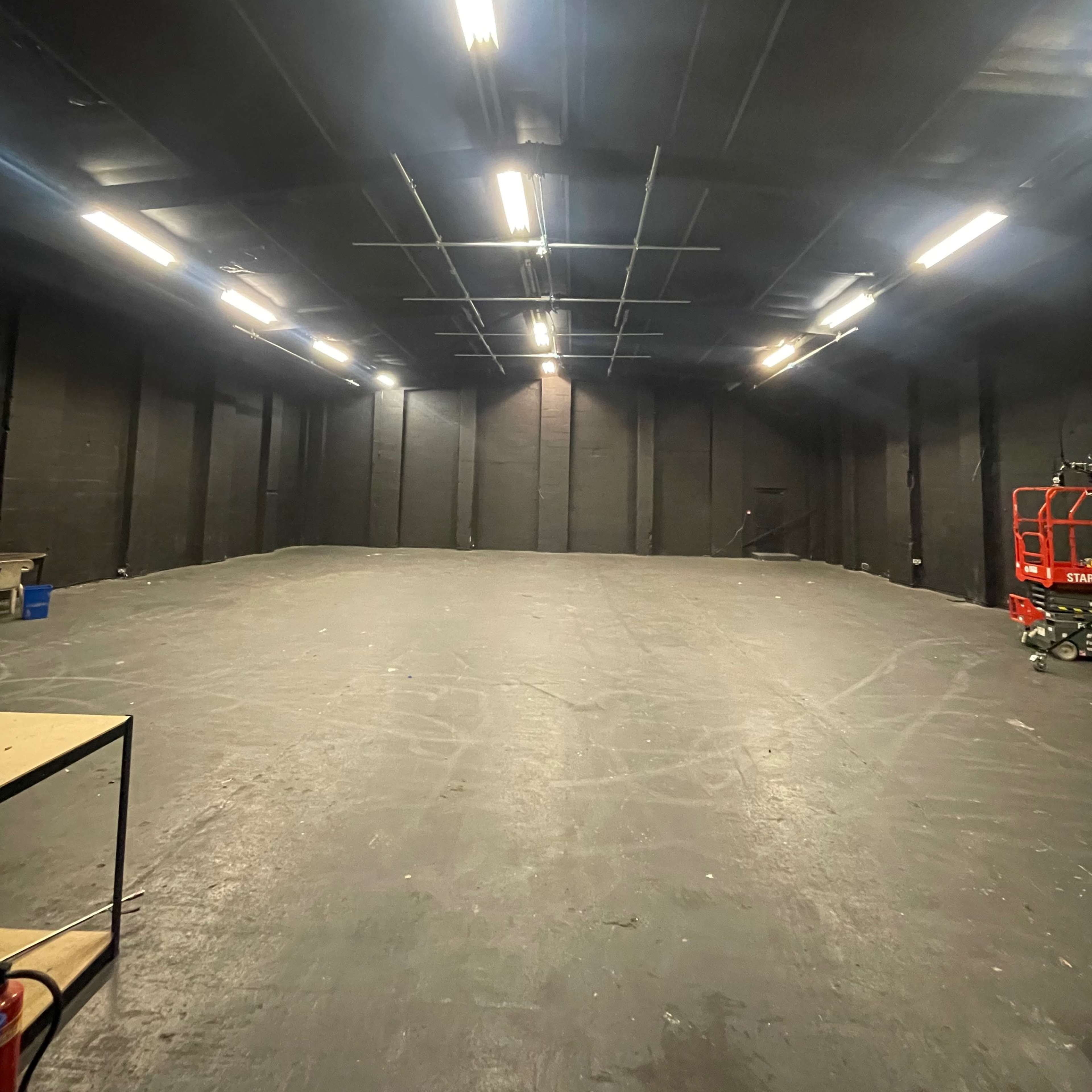 An empty, dimly lit industrial space with black walls and a smooth floor, featuring overhead lights and a small table on the left.