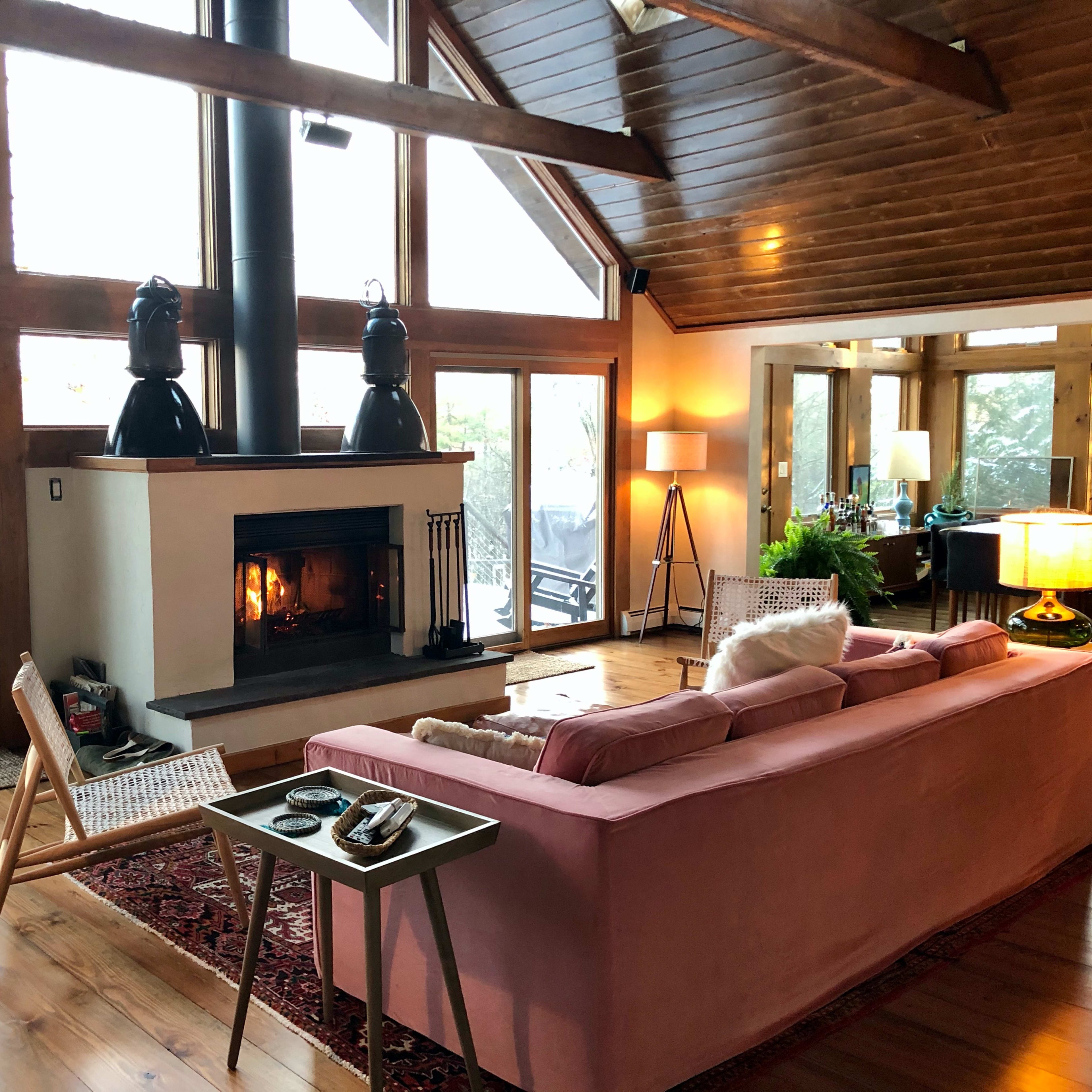 A cozy living room with a pink sofa facing a fireplace, large windows letting in natural light, and wooden beams on the ceiling.