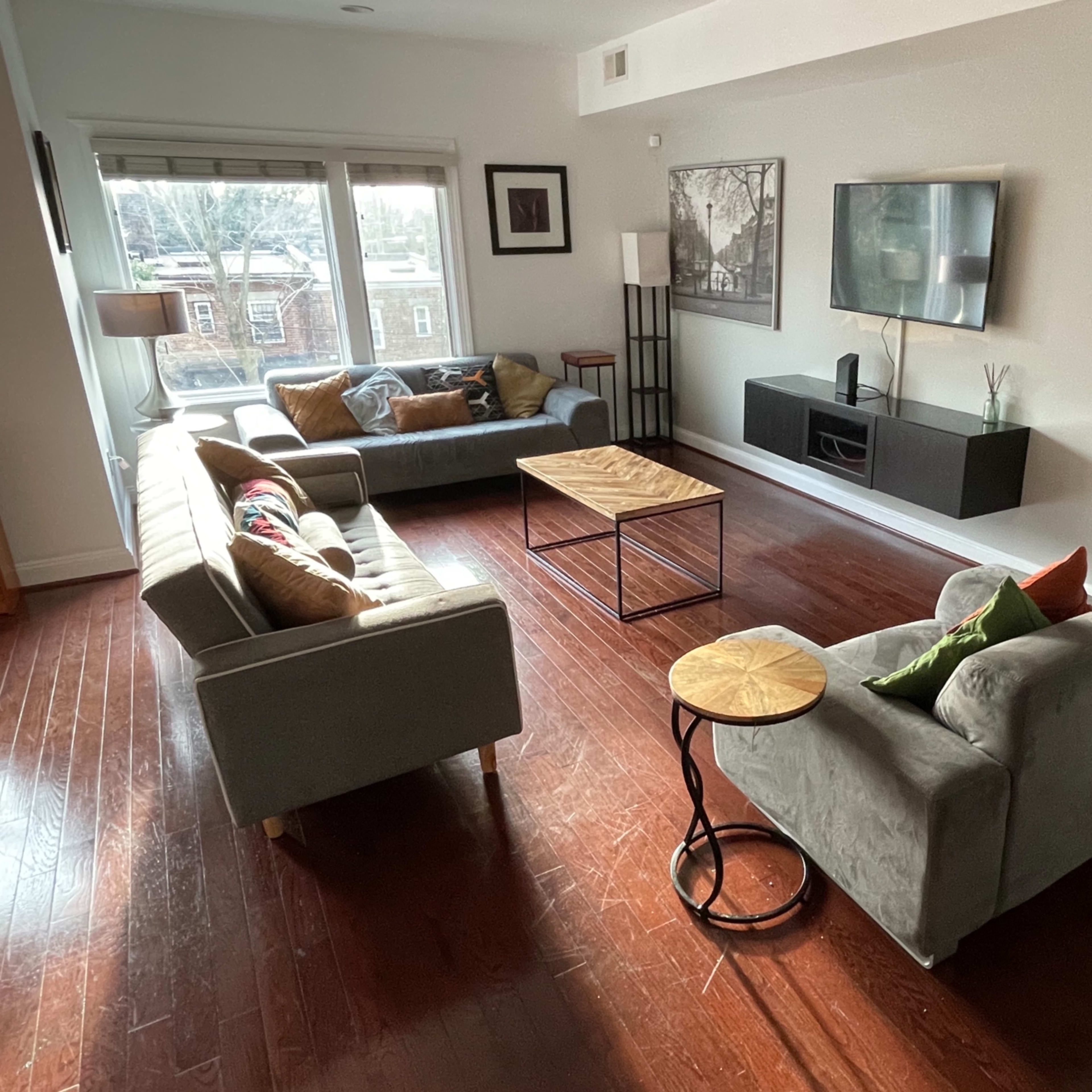 A living room features two sofas, a coffee table, and artwork on the walls, with large windows allowing natural light to enter the space.