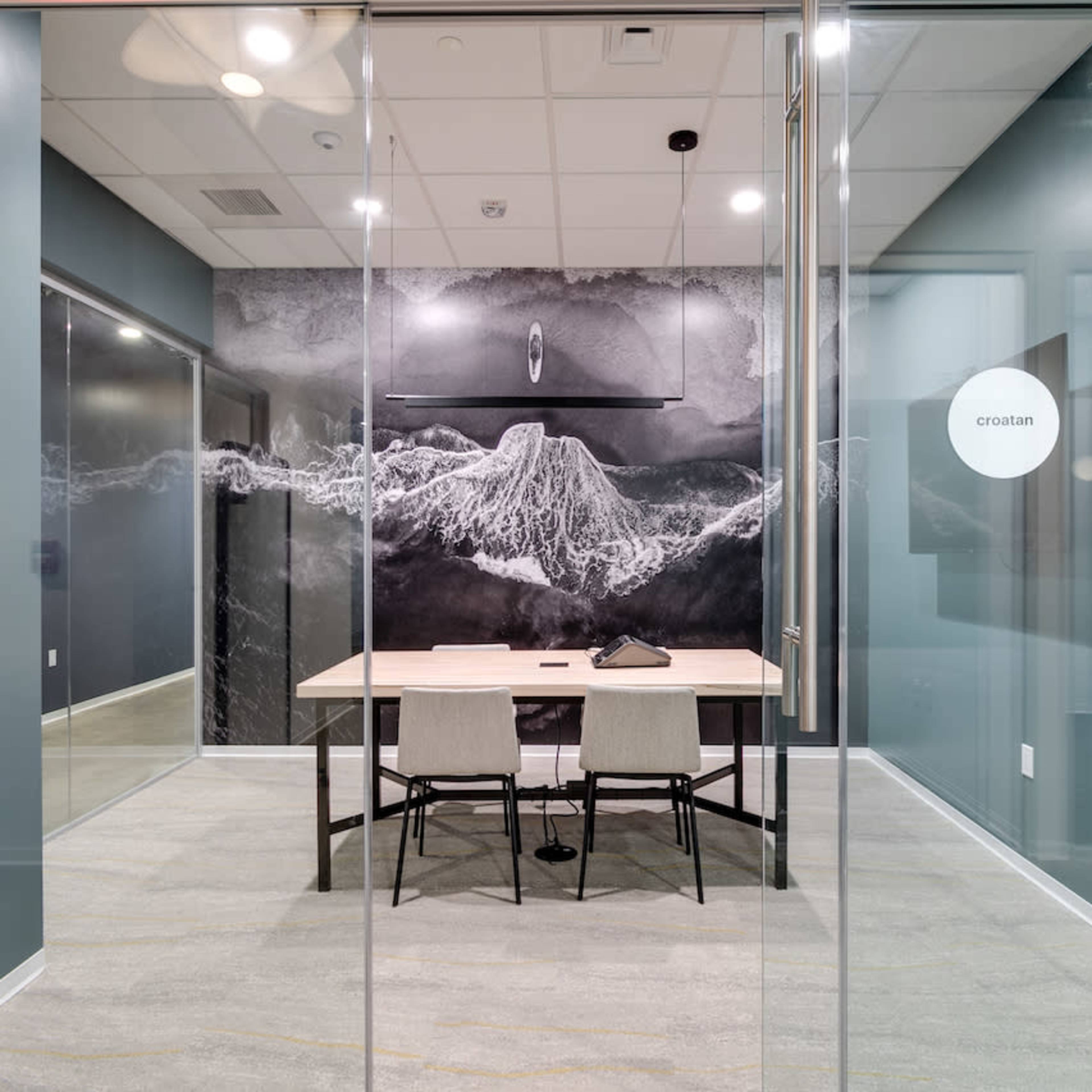 A modern conference room with a table and chairs, featuring a large mural of a mountain landscape on the wall and glass partitions.