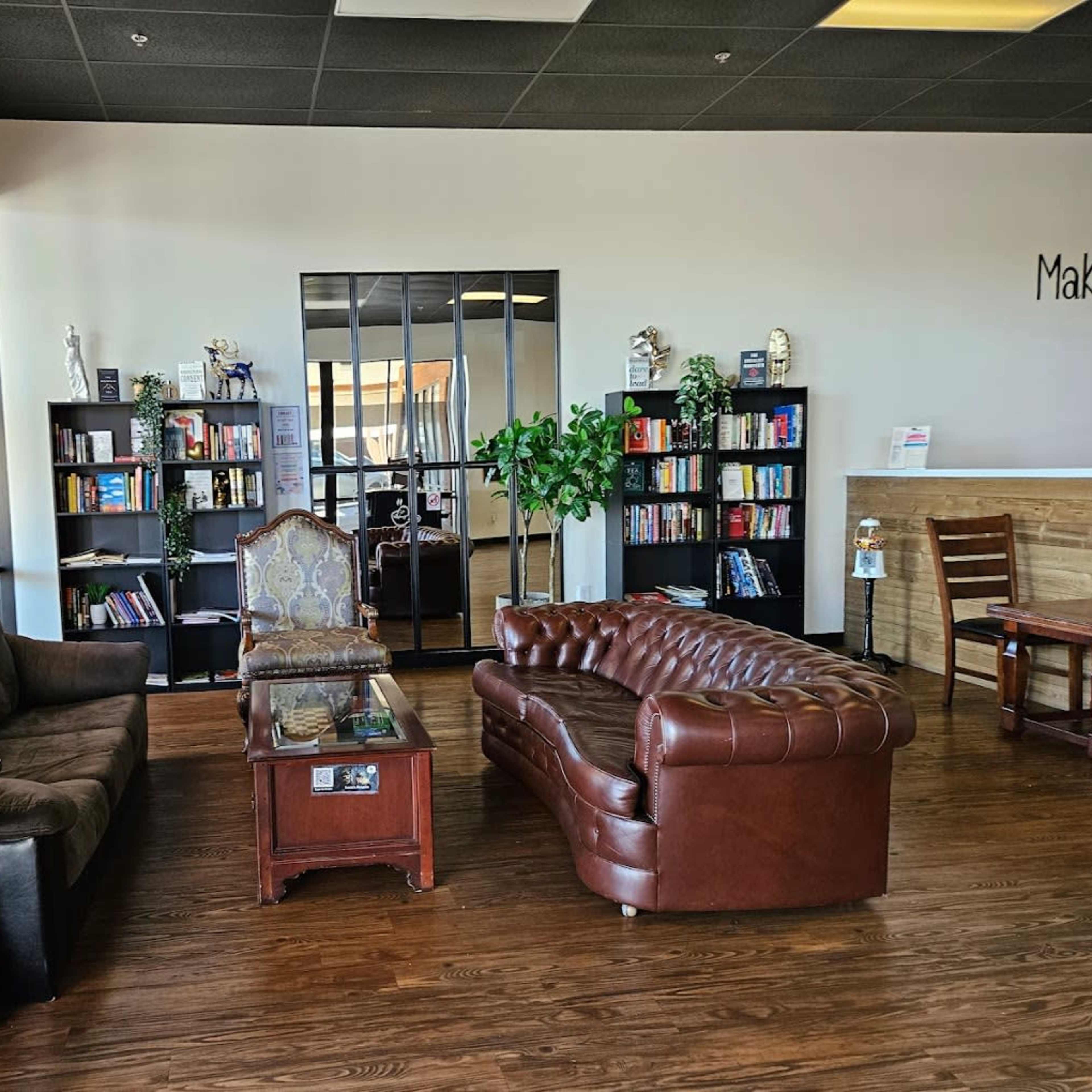 A cozy waiting area with a brown leather sofa, an ornate chair, a wooden coffee table, and bookshelves filled with books, framed by large windows allowing natural light.