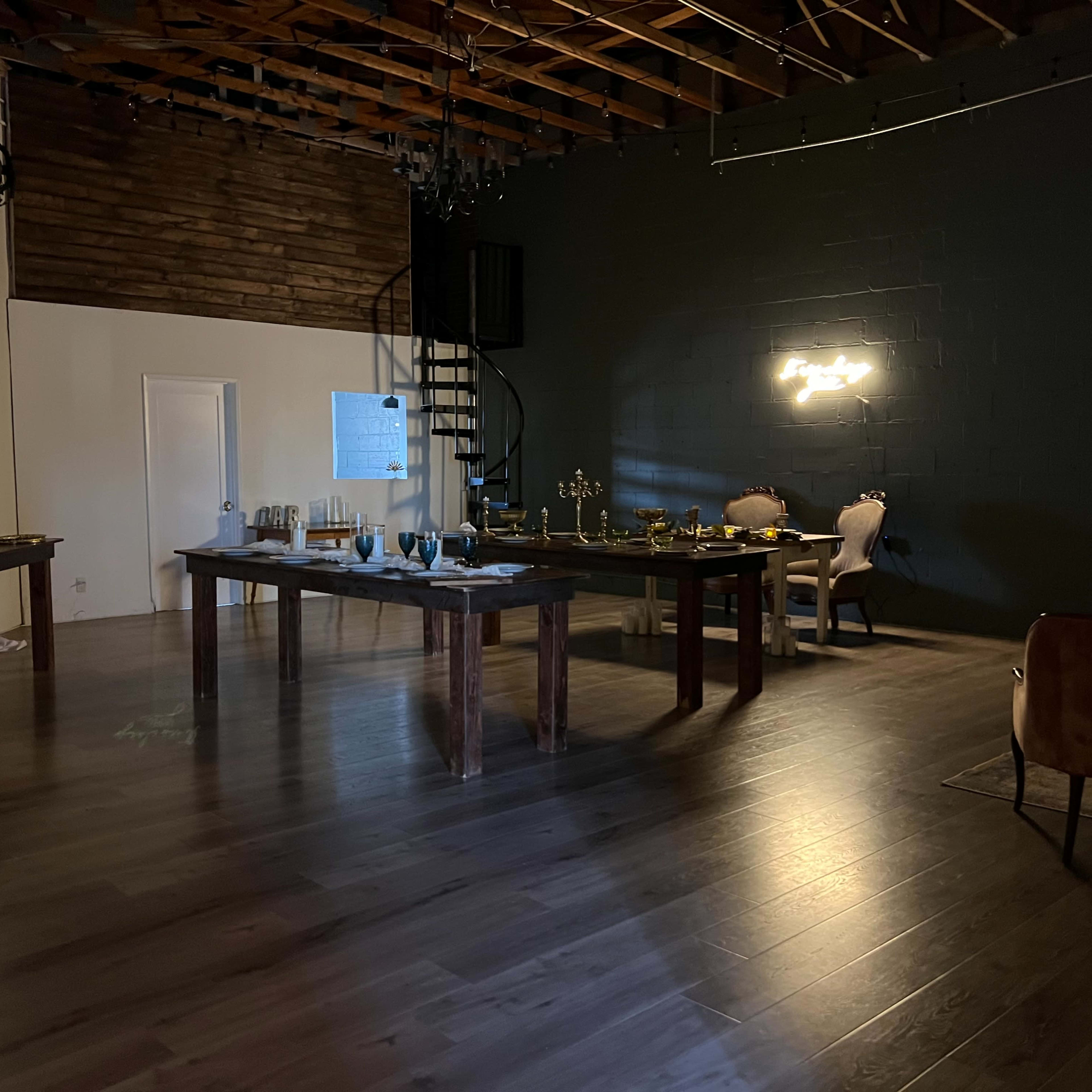 A spacious, dimly lit room with wooden tables arranged for an event, a staircase in the background, and decorative lighting on the walls.