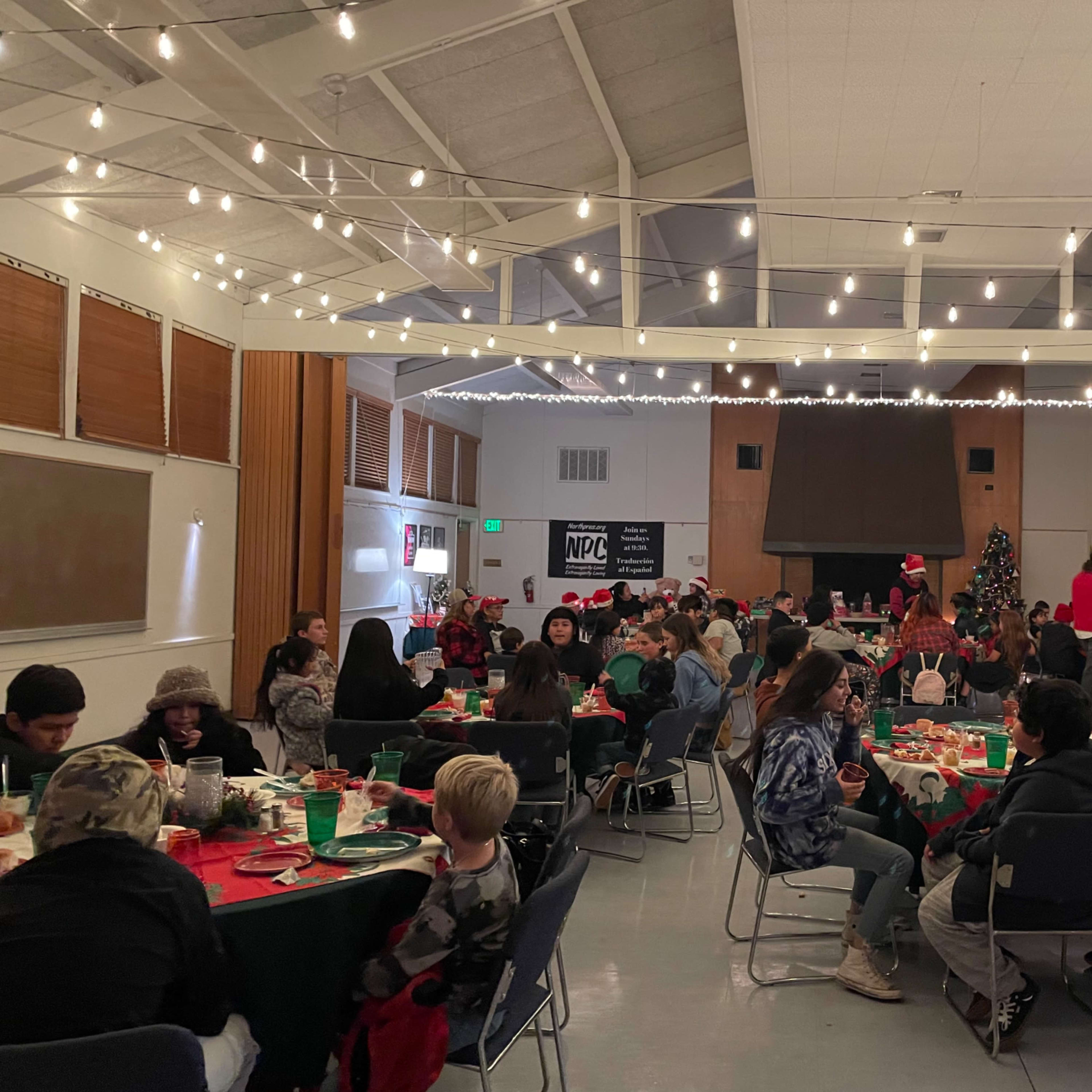 A large indoor space is filled with tables of people gathering for a holiday event, decorated with string lights and a Christmas tree.
