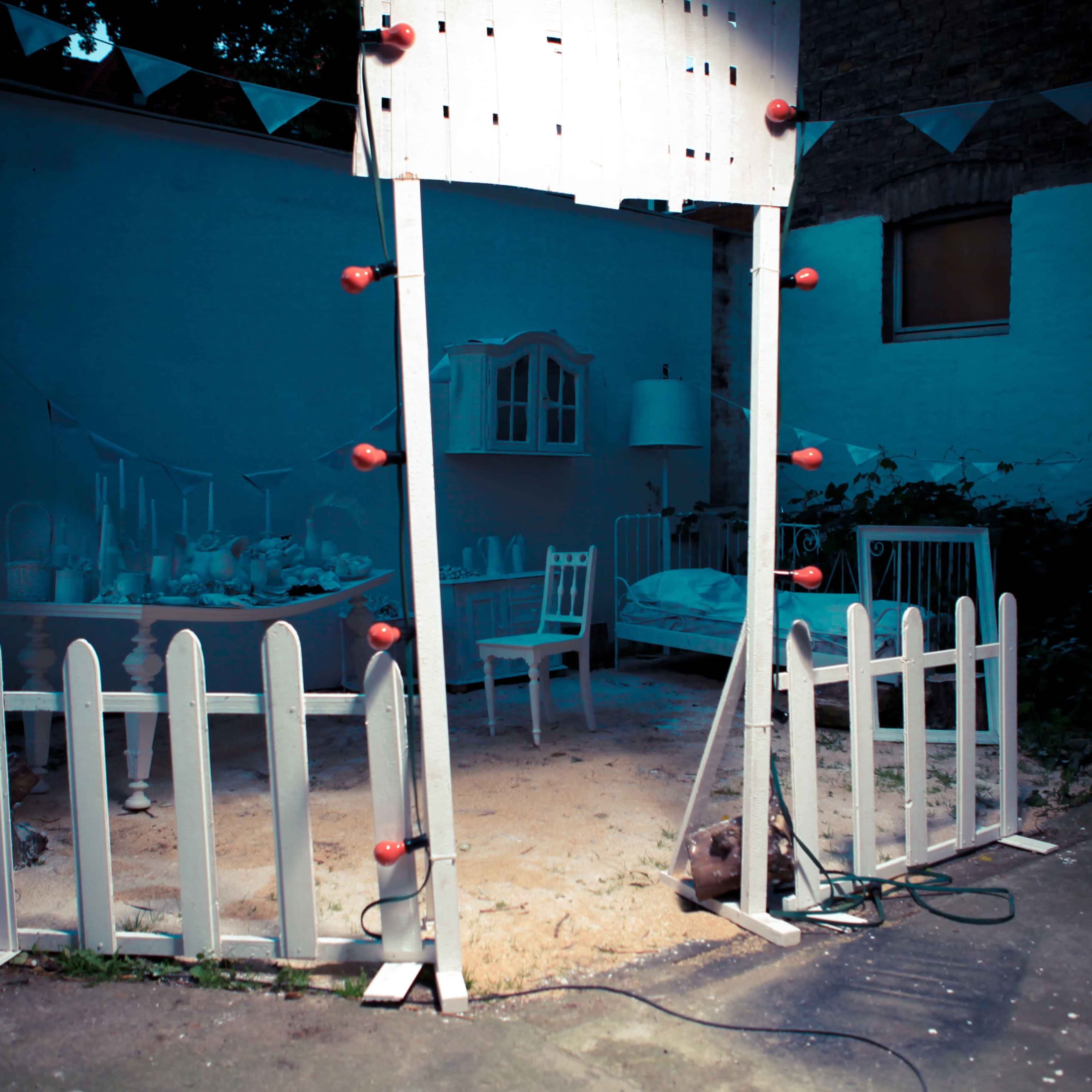 A lighted wooden archway leading to a sandy area surrounded by white picket fencing, with a sparse arrangement of furniture in the background.