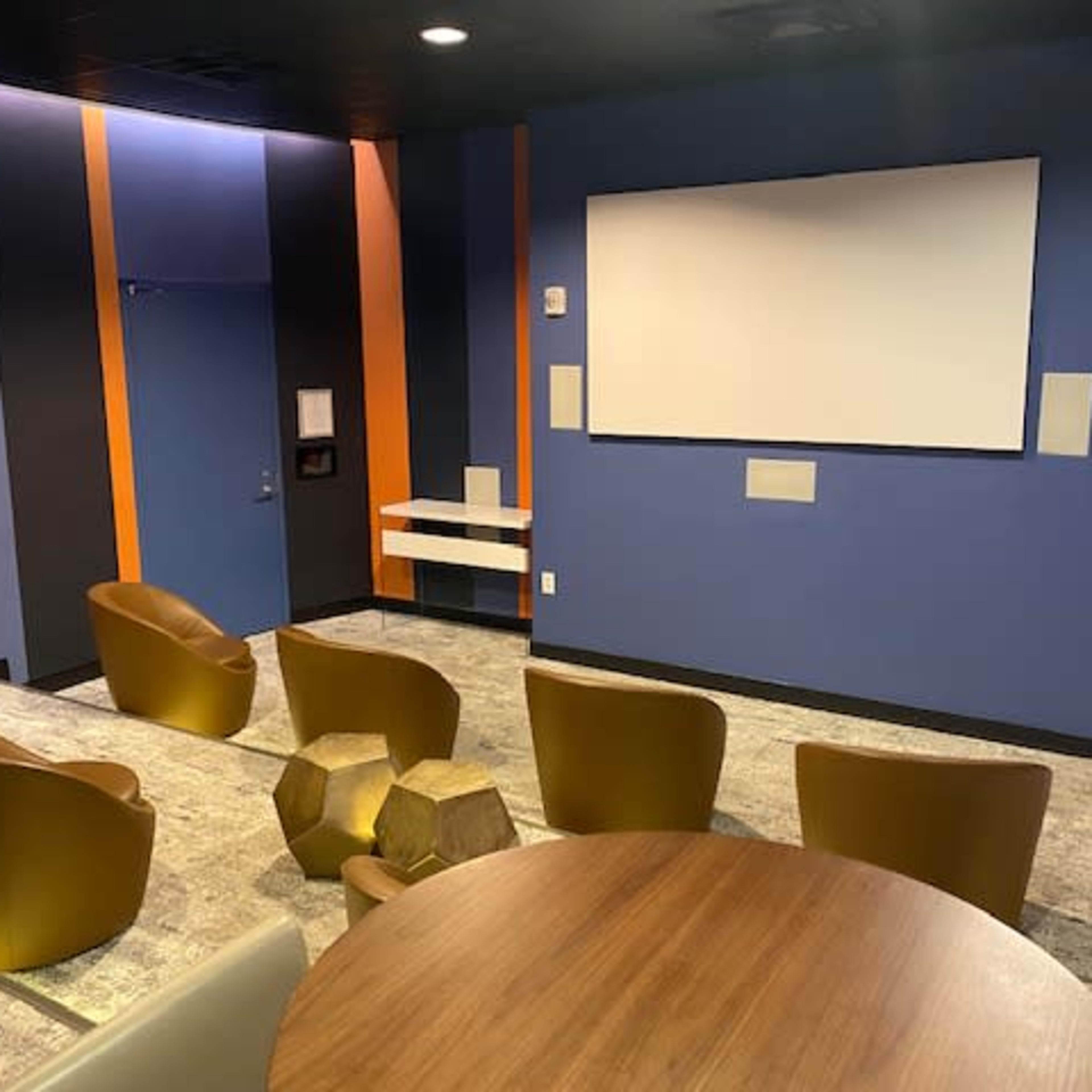 A small theater room with colorful walls, a large screen at the front, and several gold chairs arranged in a semi-circle around a wooden table.