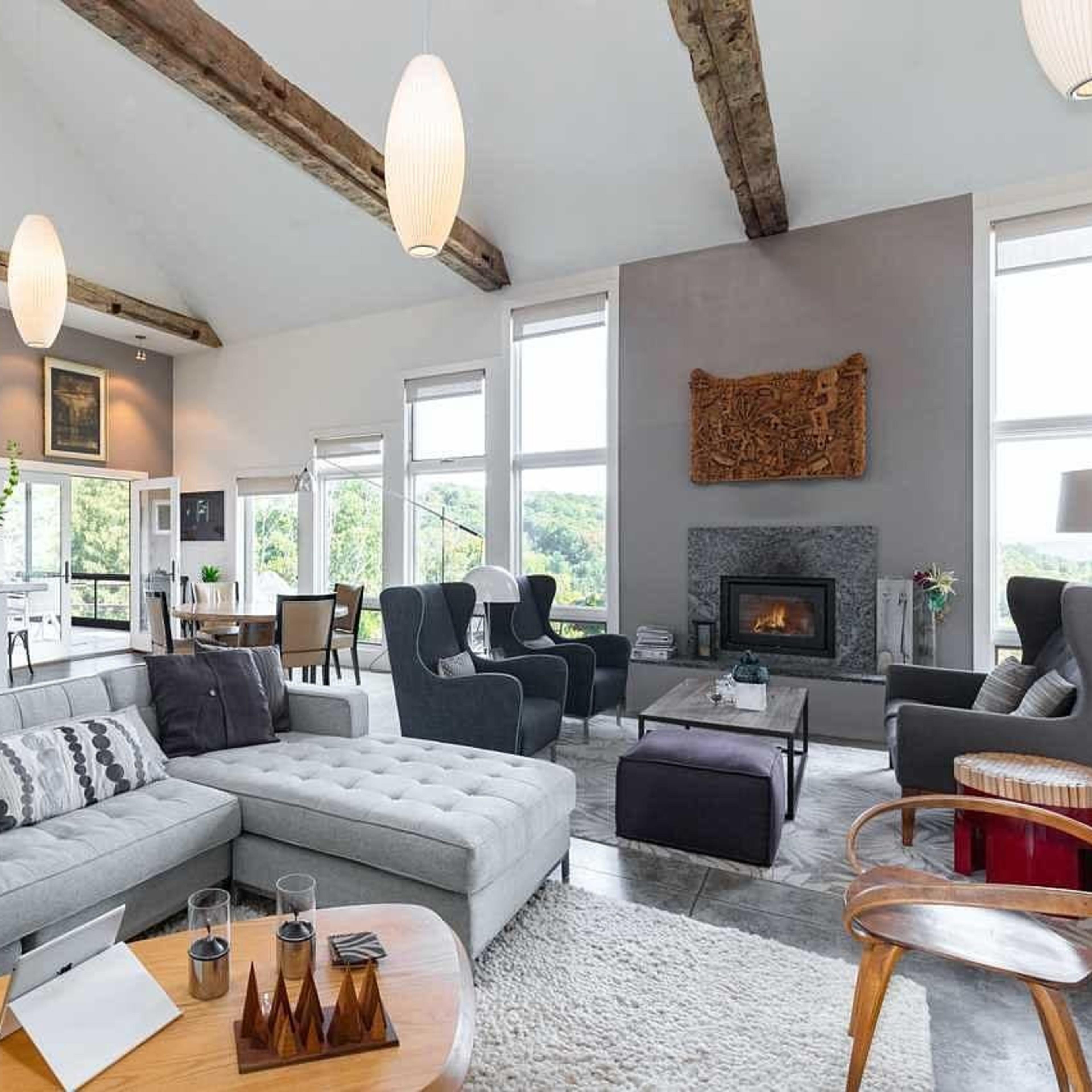 A spacious living area features a gray sectional sofa, wooden beams, and large windows with a view of the outdoors.