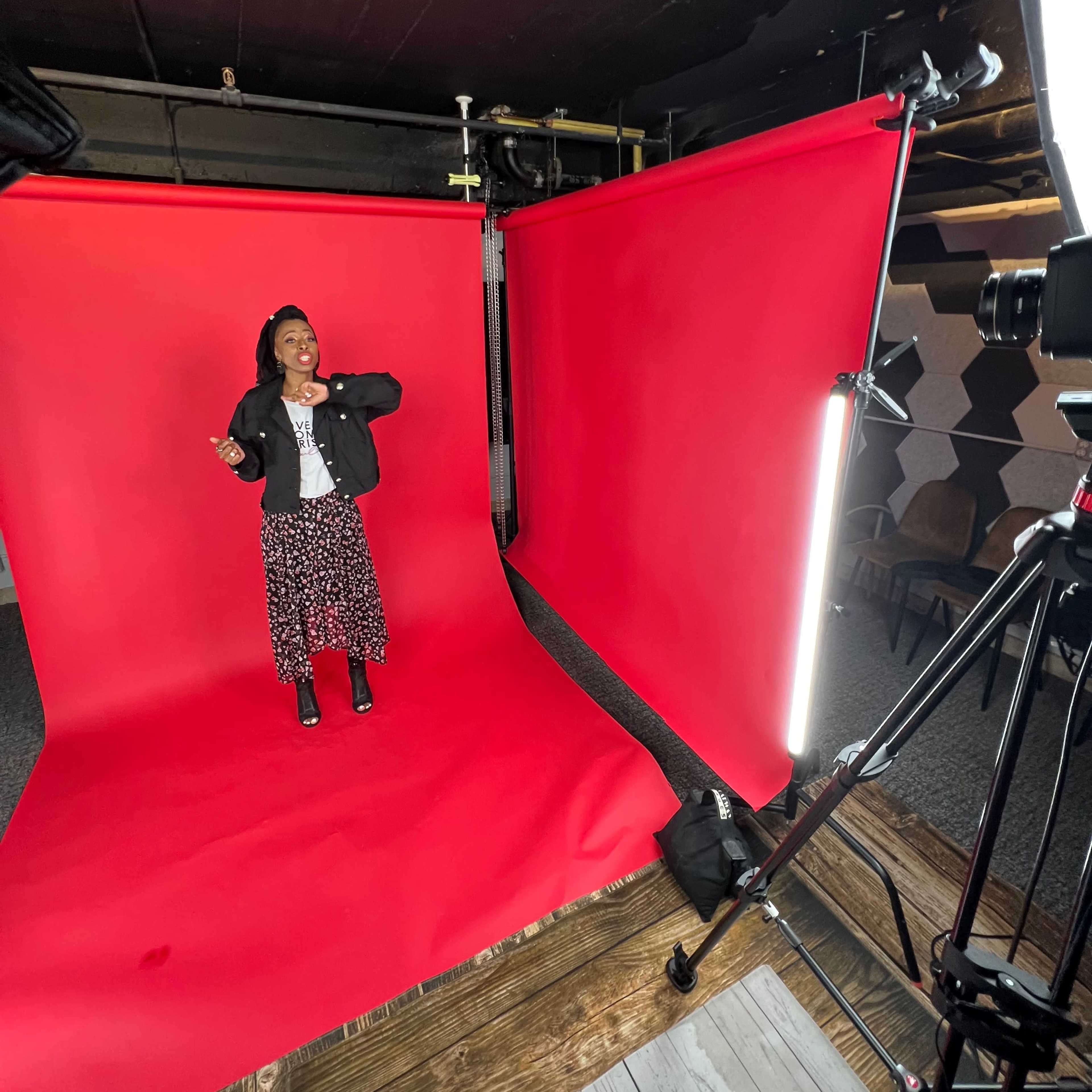 A person stands in front of a vibrant red backdrop in a photography studio, with studio lights and a camera set up nearby.