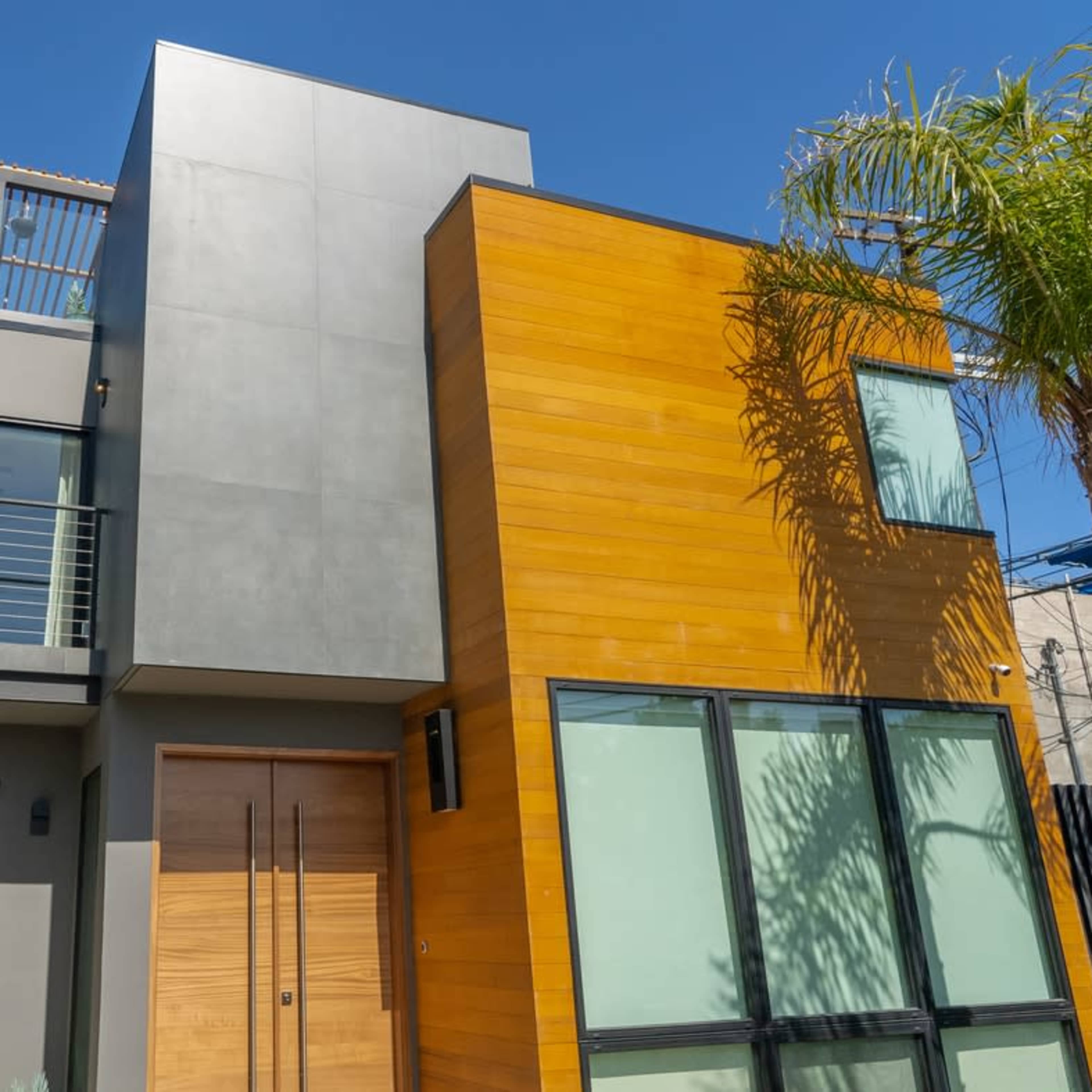 A modern two-story house with a combination of gray and wooden exterior walls, large windows, and a rooftop terrace.