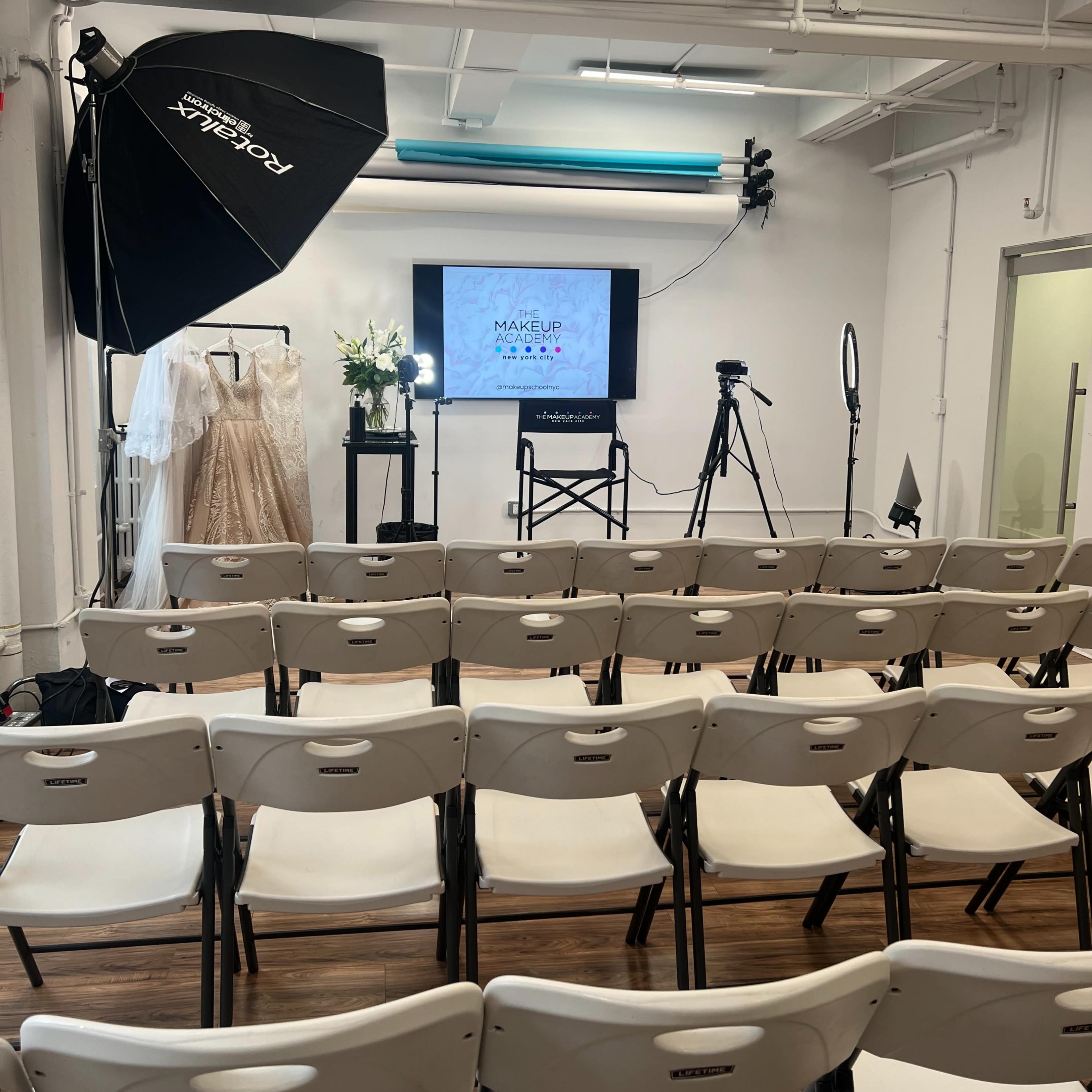A spacious room set up for a makeup workshop, featuring rows of white folding chairs, a screen displaying a makeup event title, and various filming equipment.