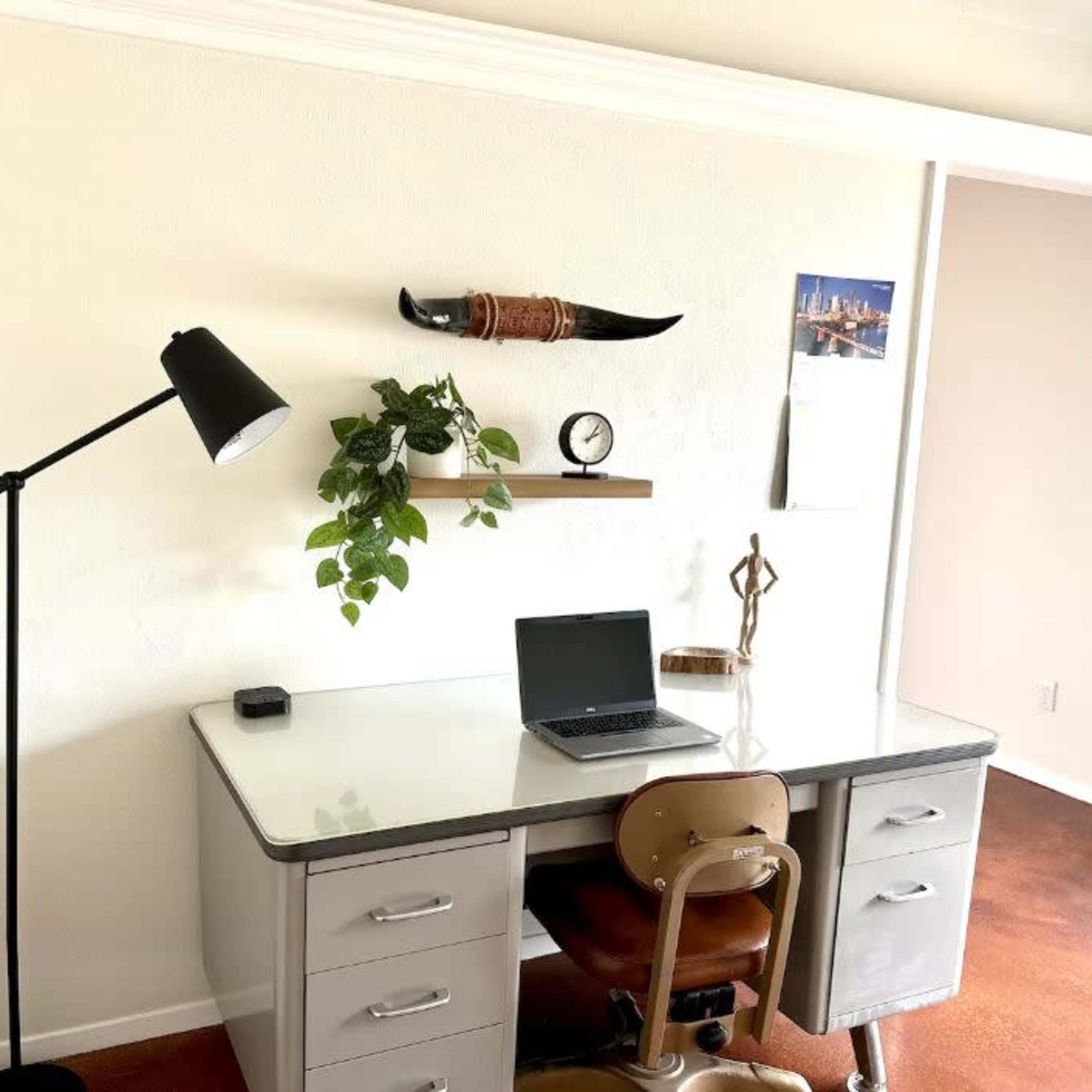 A minimalist workspace features a desk with a laptop, a plant on a shelf, a decorative item, and a standing lamp against a light-colored wall.