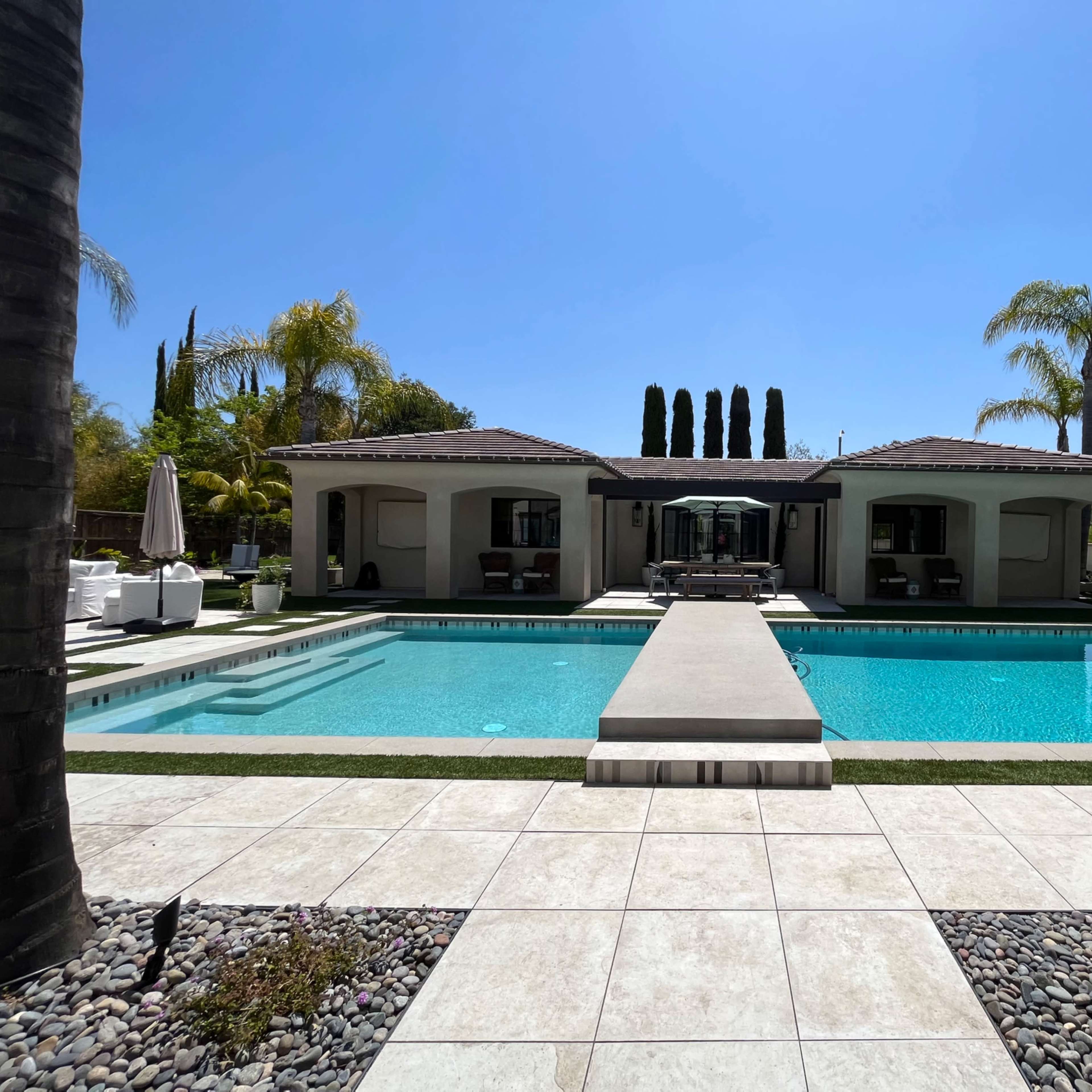 A rectangular swimming pool is bordered by a stone patio and leads to a single-story house with multiple palm trees in the background.