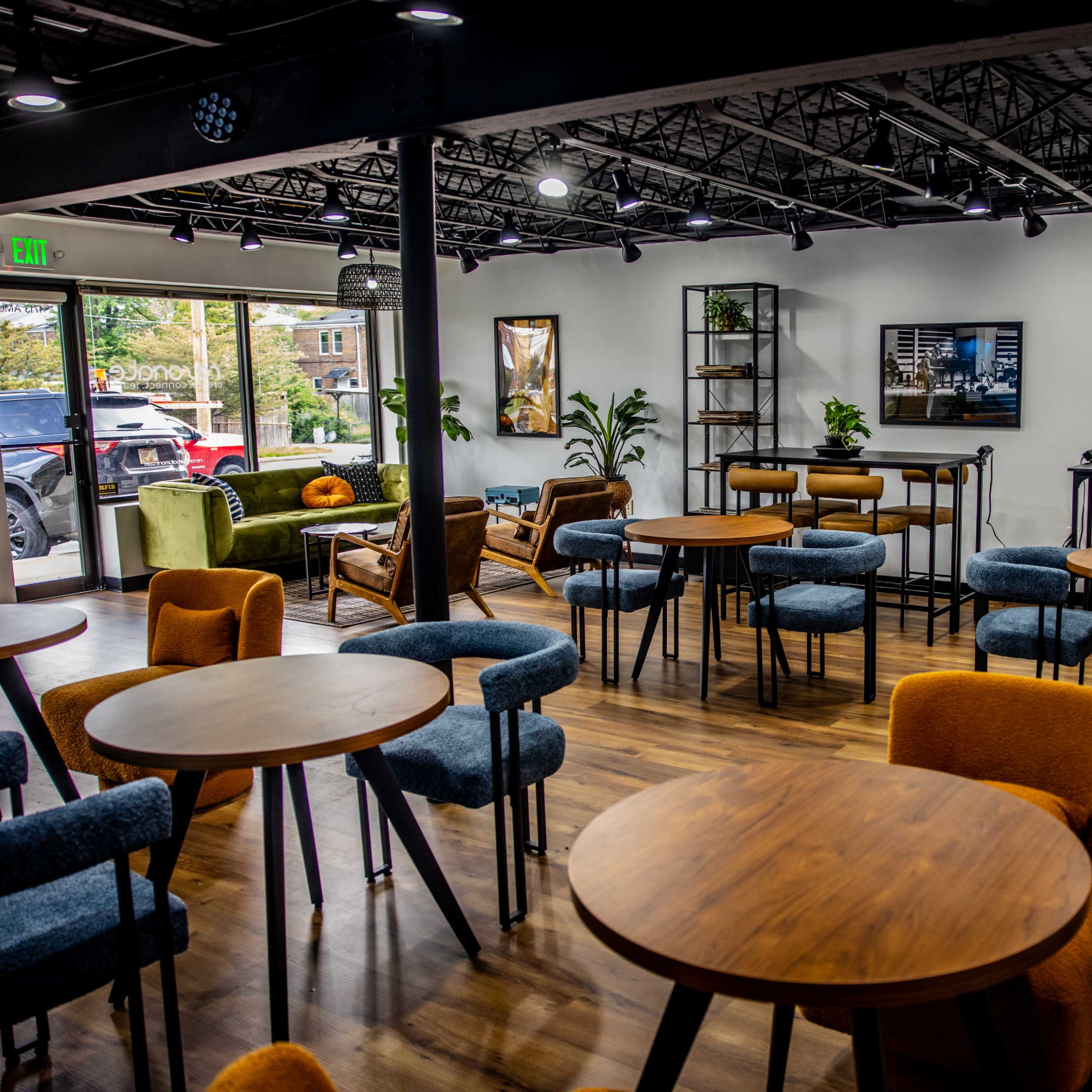A modern café interior with wooden tables, colorful chairs, and greenery, featuring large windows that allow natural light to enter.