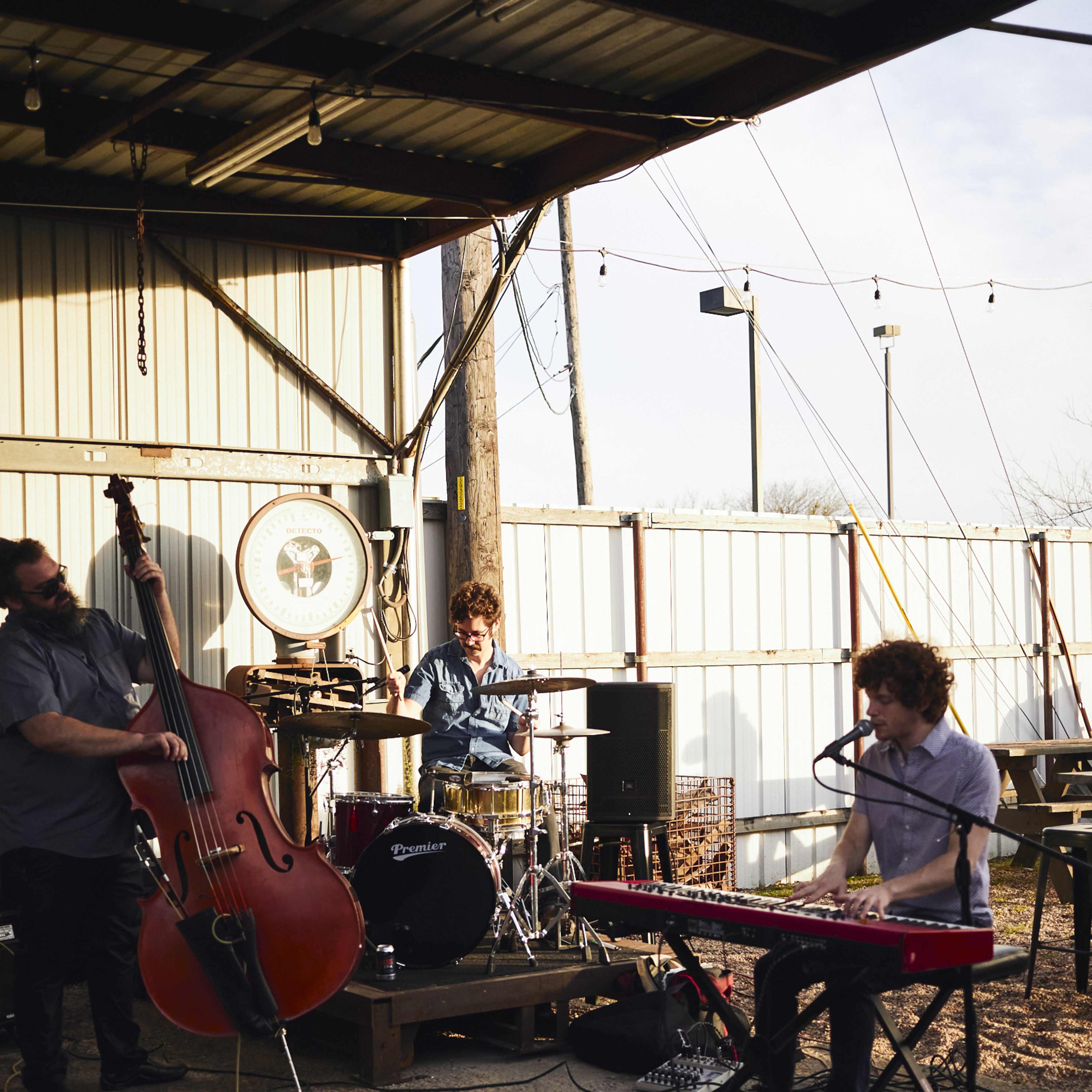 Three musicians perform with a double bass, drums, and a keyboard in an outdoor venue under a metal roof.