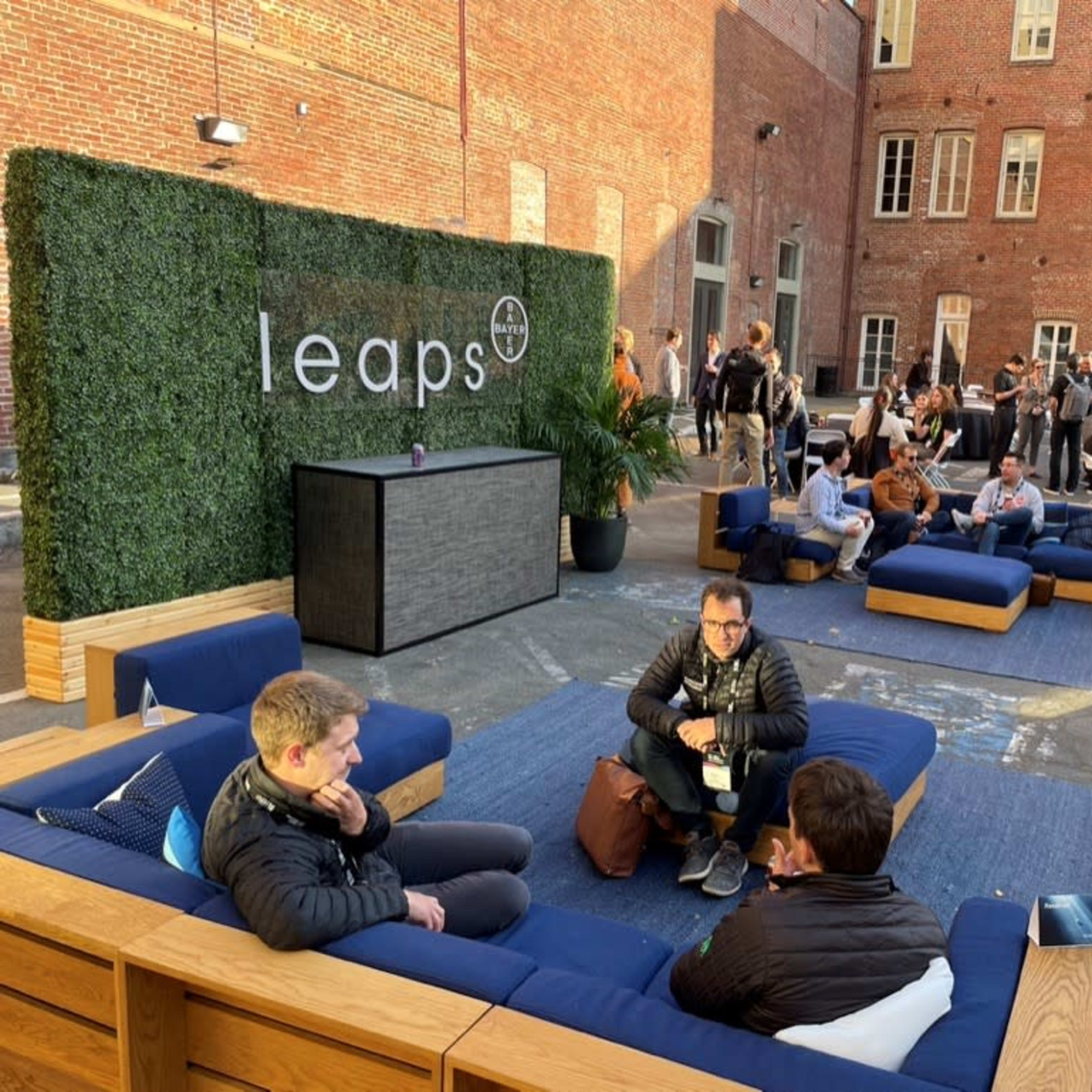 A courtyard features modern seating arrangements with attendees engaged in conversation, surrounded by greenery and a backdrop of brick buildings.