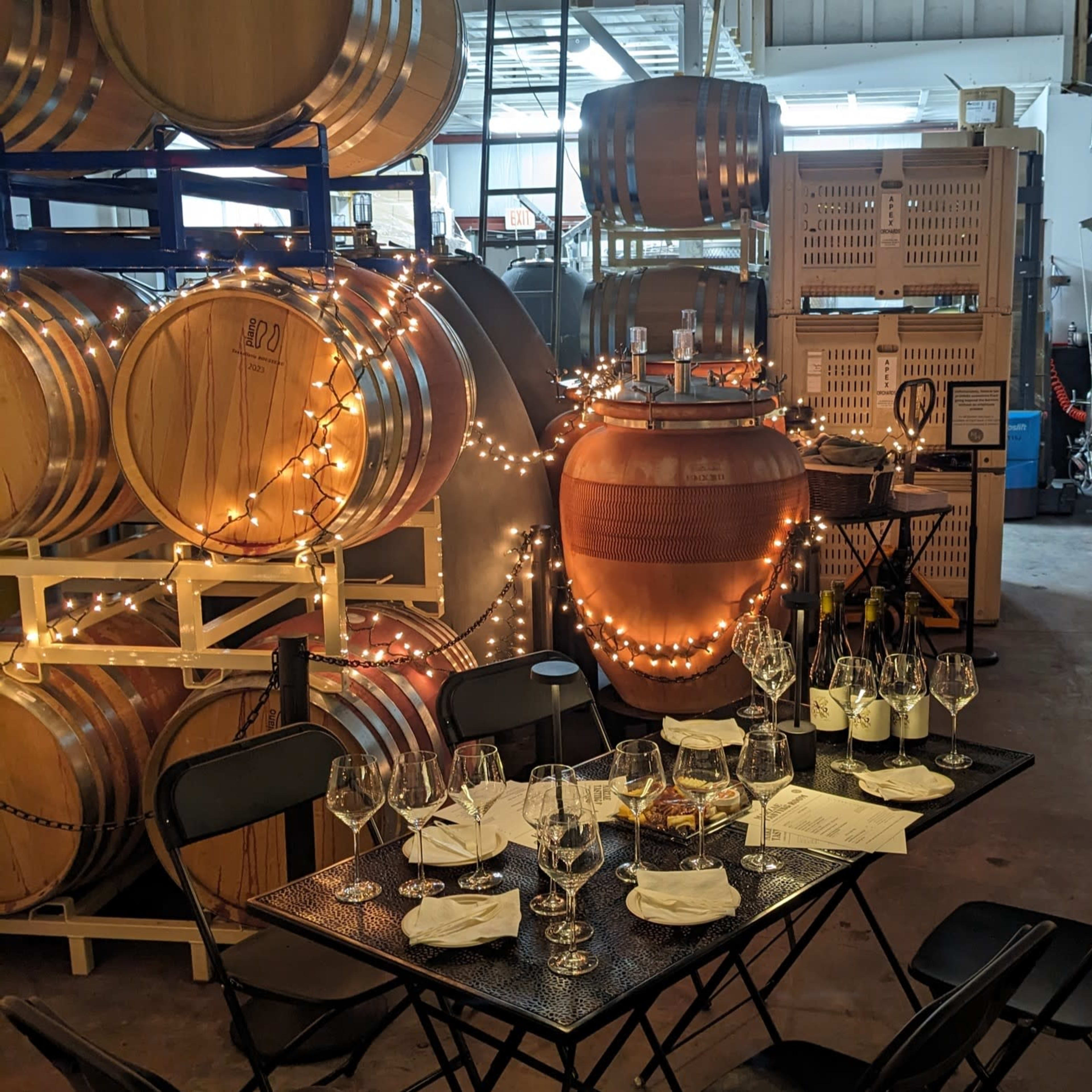 A table set for a wine tasting is positioned among barrels and fermentation tanks in a winery.