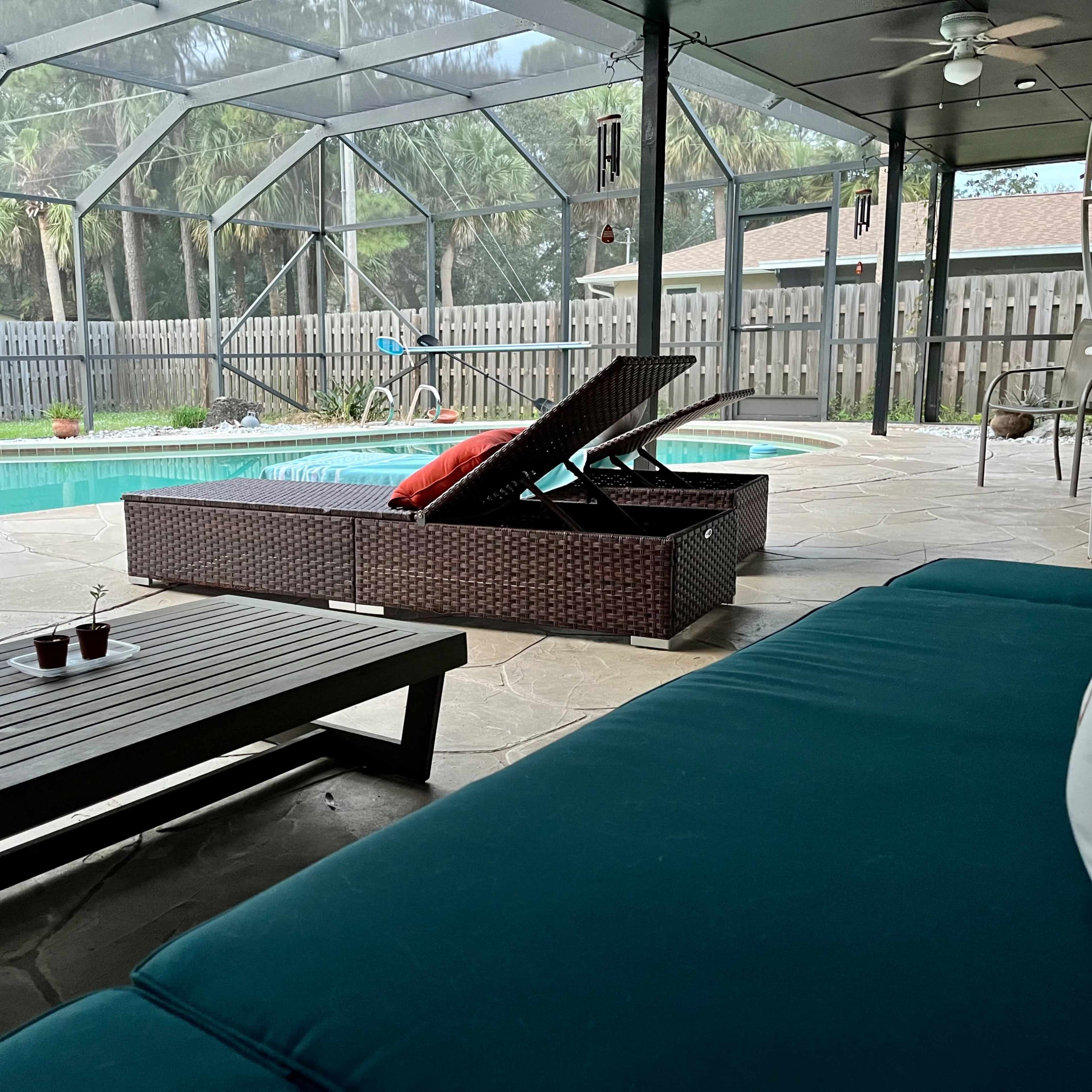 A screened-in patio with a swimming pool, a lounge chair, a table, and surrounding palm trees.