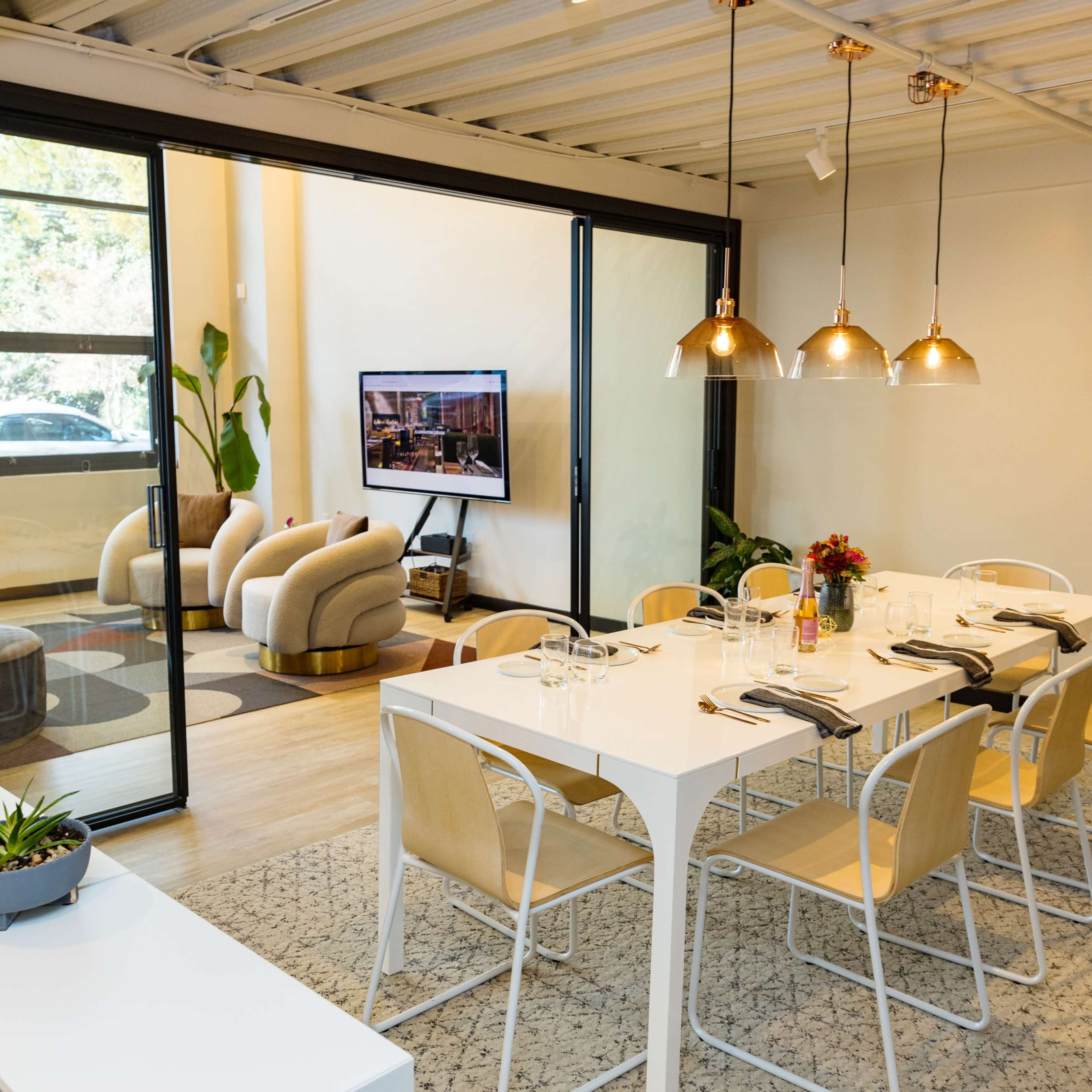 A modern dining area with a long white table set for a meal, alongside a cozy seating area visible through a glass partition.