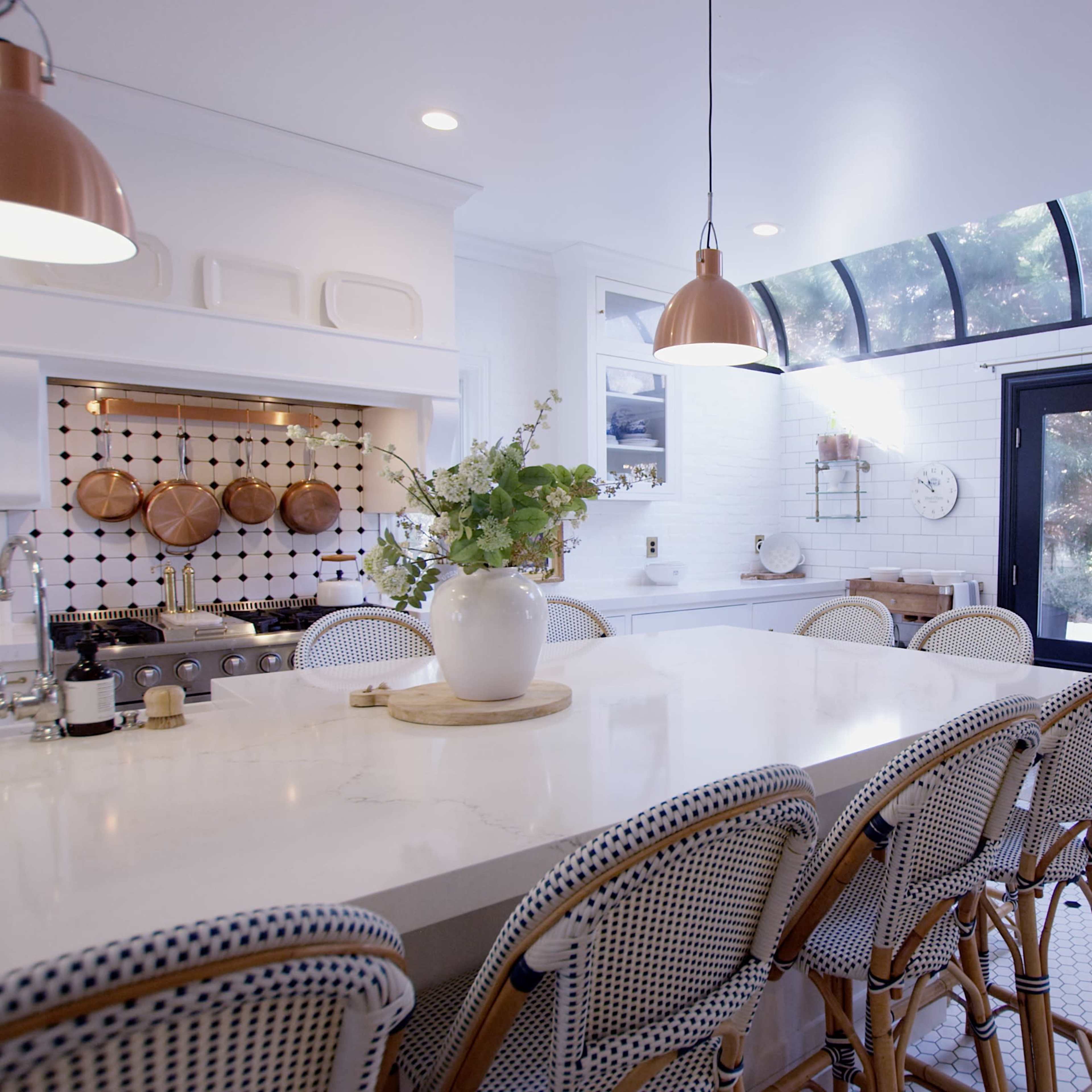 A spacious kitchen features an island with high-backed chairs, wooden cookware displayed on the wall, and a large window allowing natural light to fill the space.