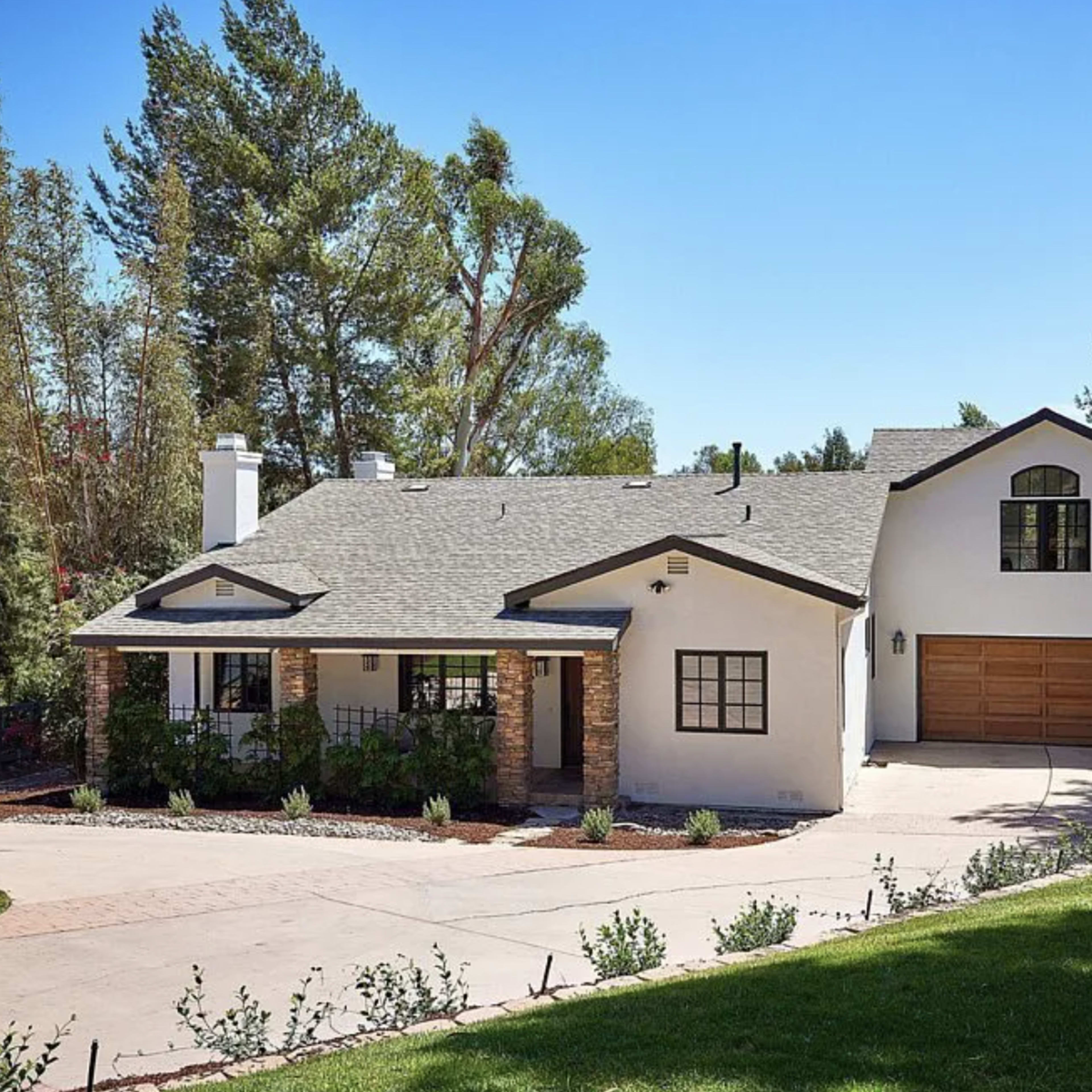 A large, two-story house with a gray roof and white exterior is situated on a landscaped lot with a circular driveway and garage.