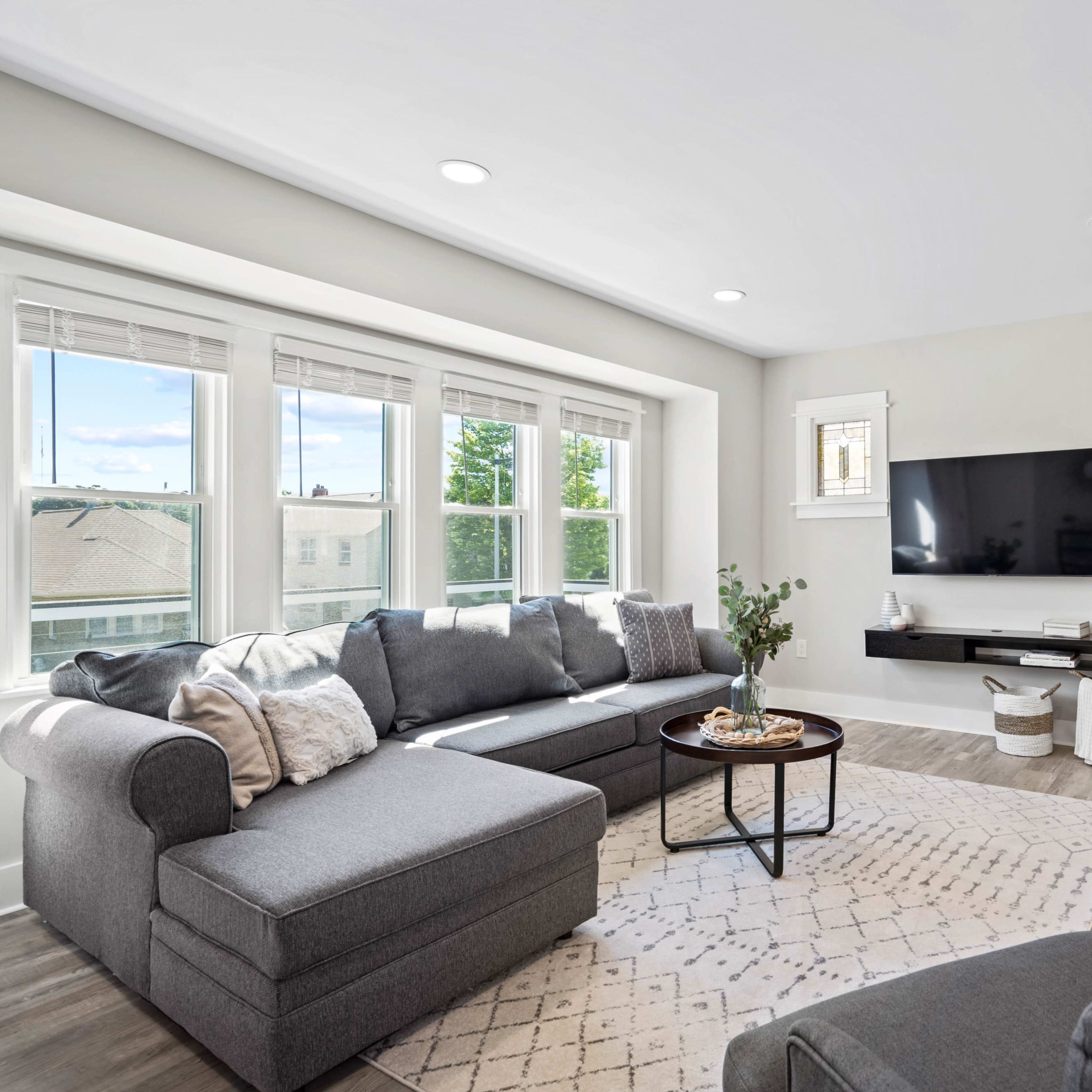 A modern living room with a sectional sofa, a coffee table, and a mounted television on a light-colored wall, illuminated by natural light from large windows.