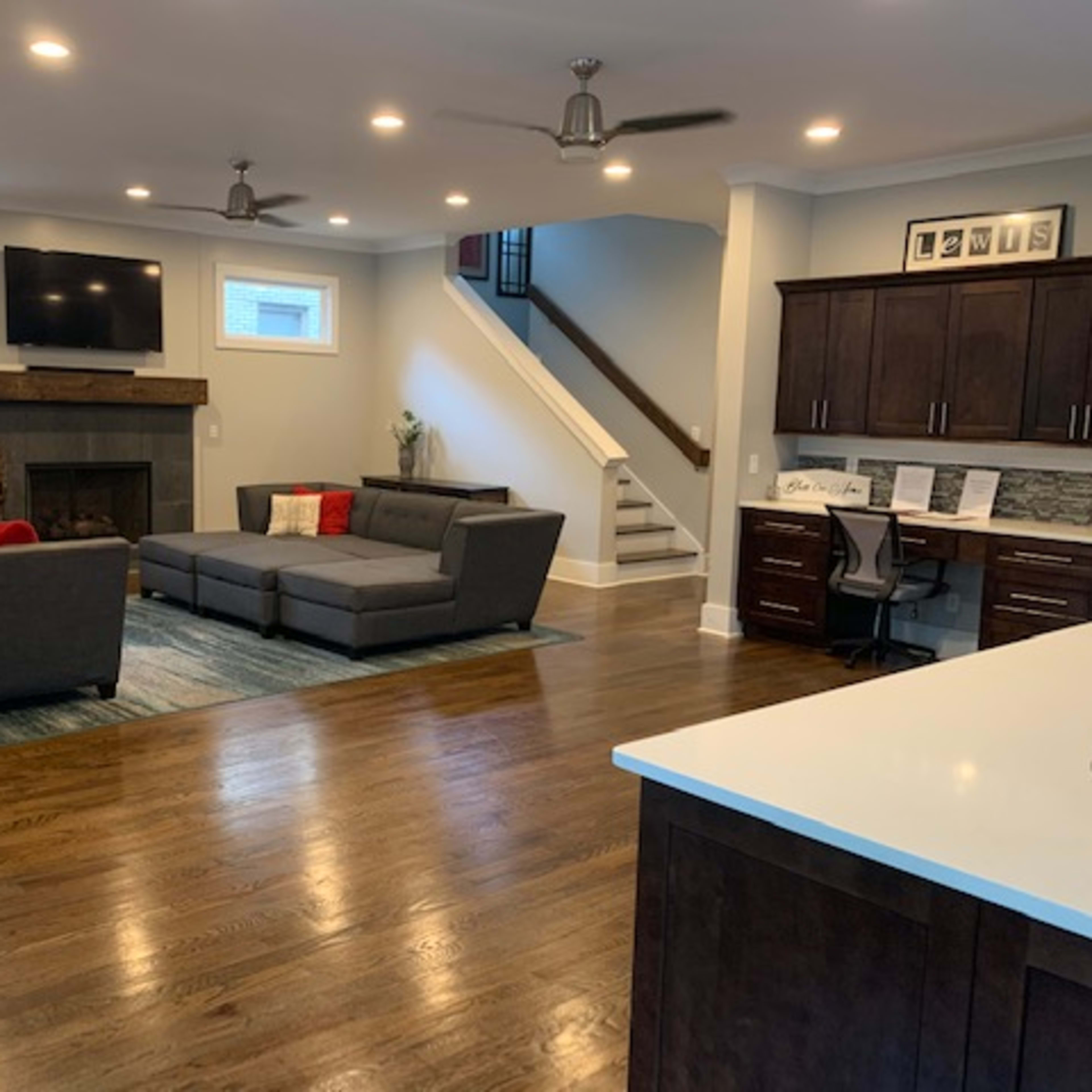 A modern living room with a gray sectional sofa, a fireplace, and a staircase leading to an upper level, all illuminated by recessed lighting.